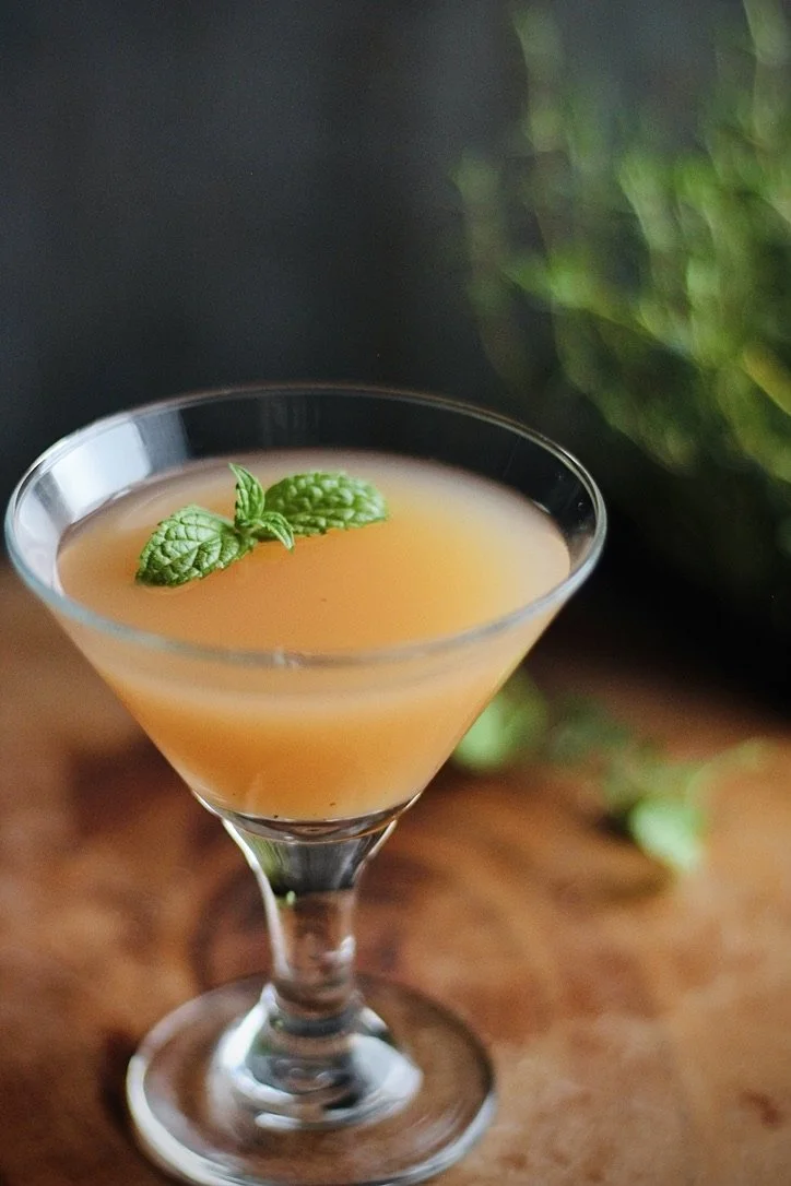 Cocktail in a martini glass garnished with a mint sprig, on a wooden surface with blurred green foliage in the background.