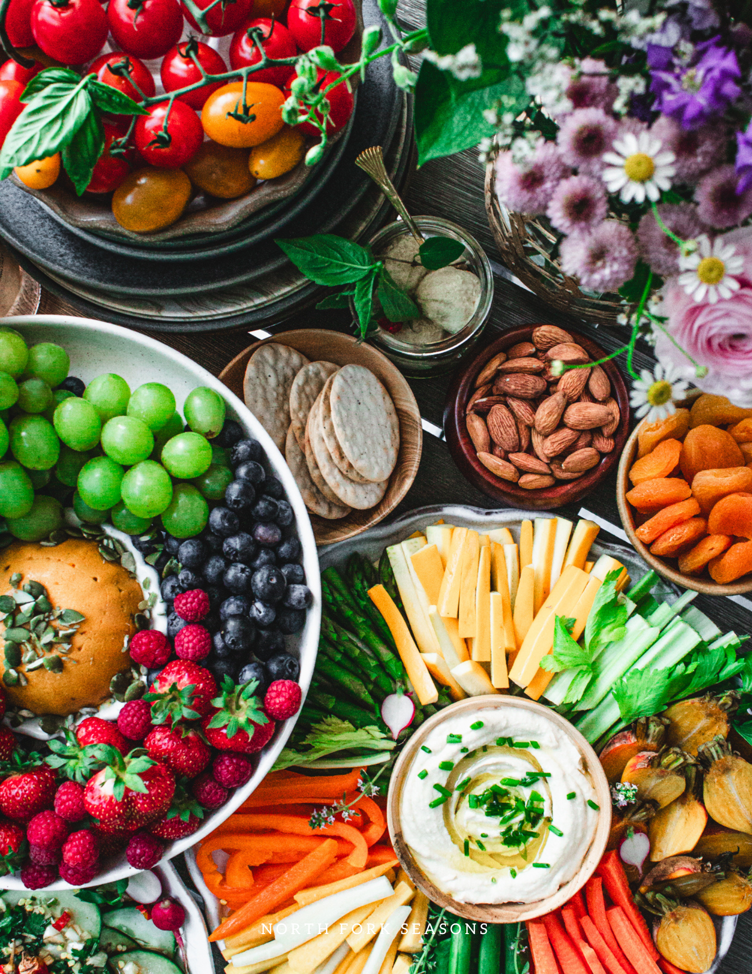 An assortment of fresh fruits, vegetables, nuts, and flowers displayed on a table, including cherry tomatoes, grapes, strawberries, raspberries, crackers, almonds, dried apricots, cheese sticks, celery, carrots, a bowl of hummus with herbs, and a bouquet of flowers.