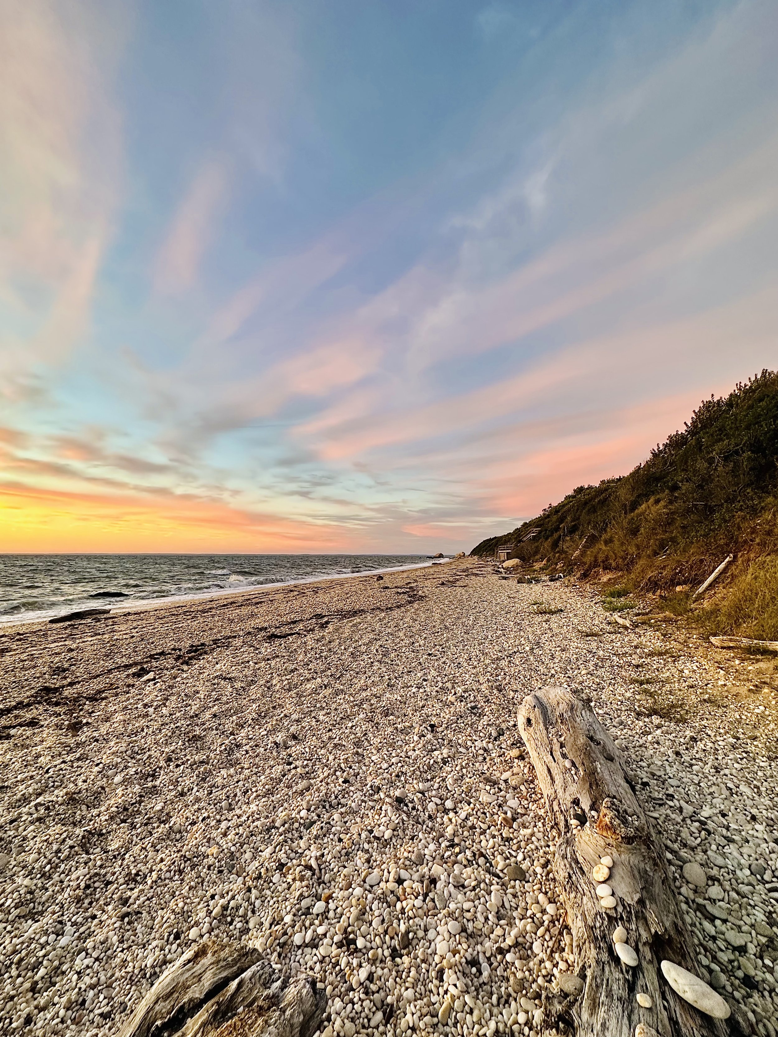 Sunset over a pebbled beach with driftwood and grassy dunes on the right.