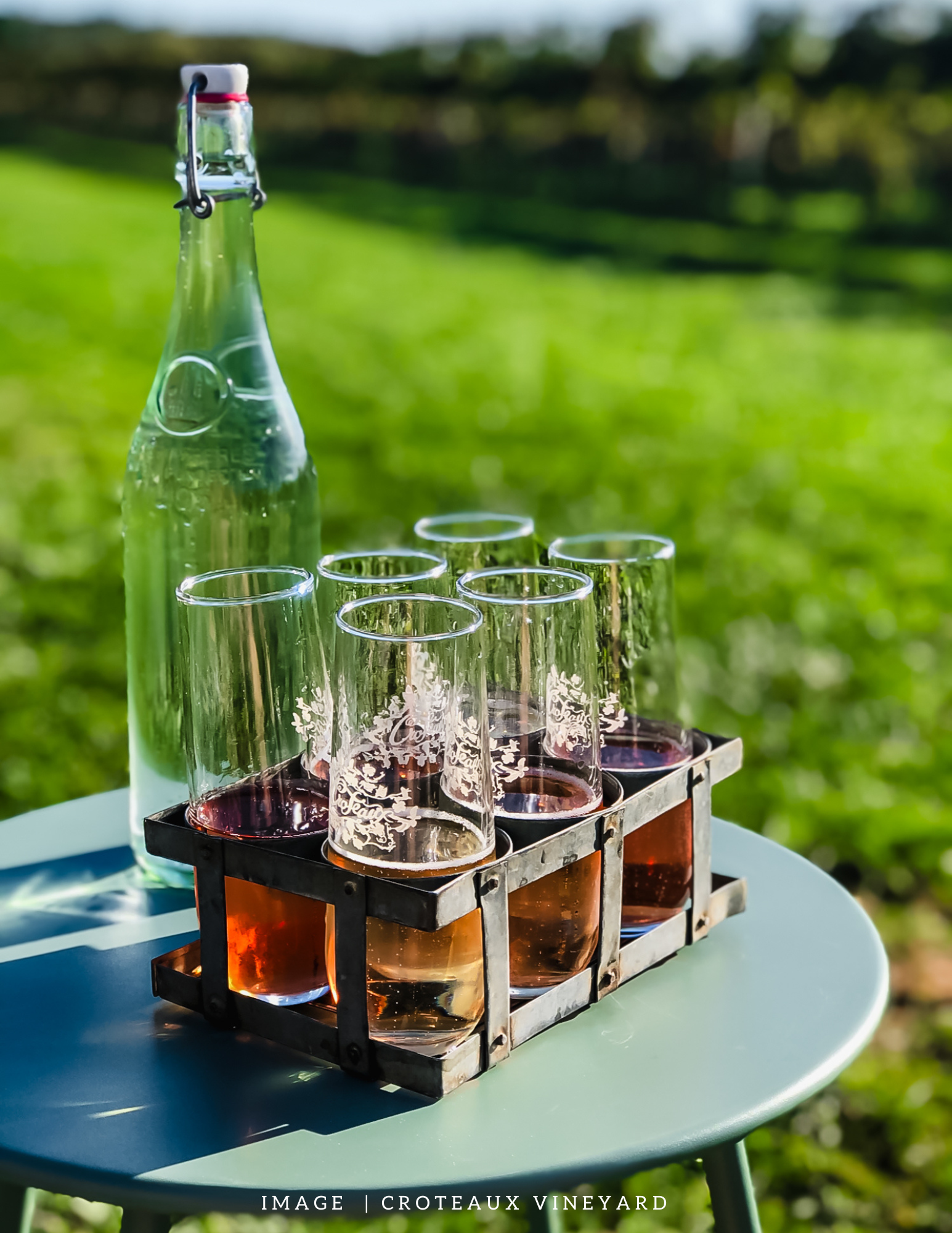 A glass bottle with a swing-top cap and six glasses filled with rosé wine on a white round table outdoors in a vineyard setting.