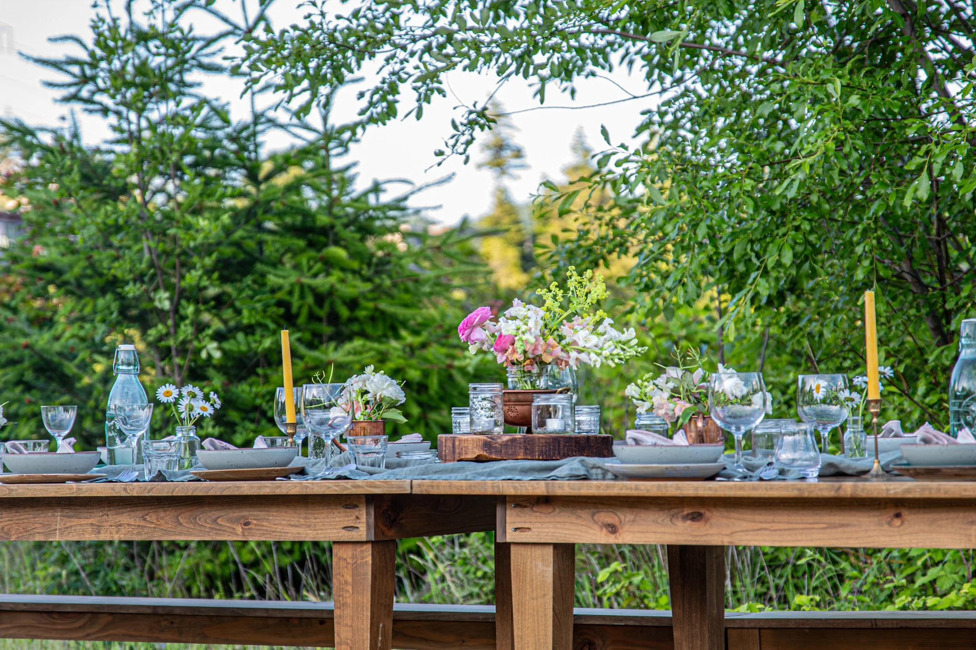 Outdoor dining table set with floral centerpieces, glassware, and candles, surrounded by lush green trees.