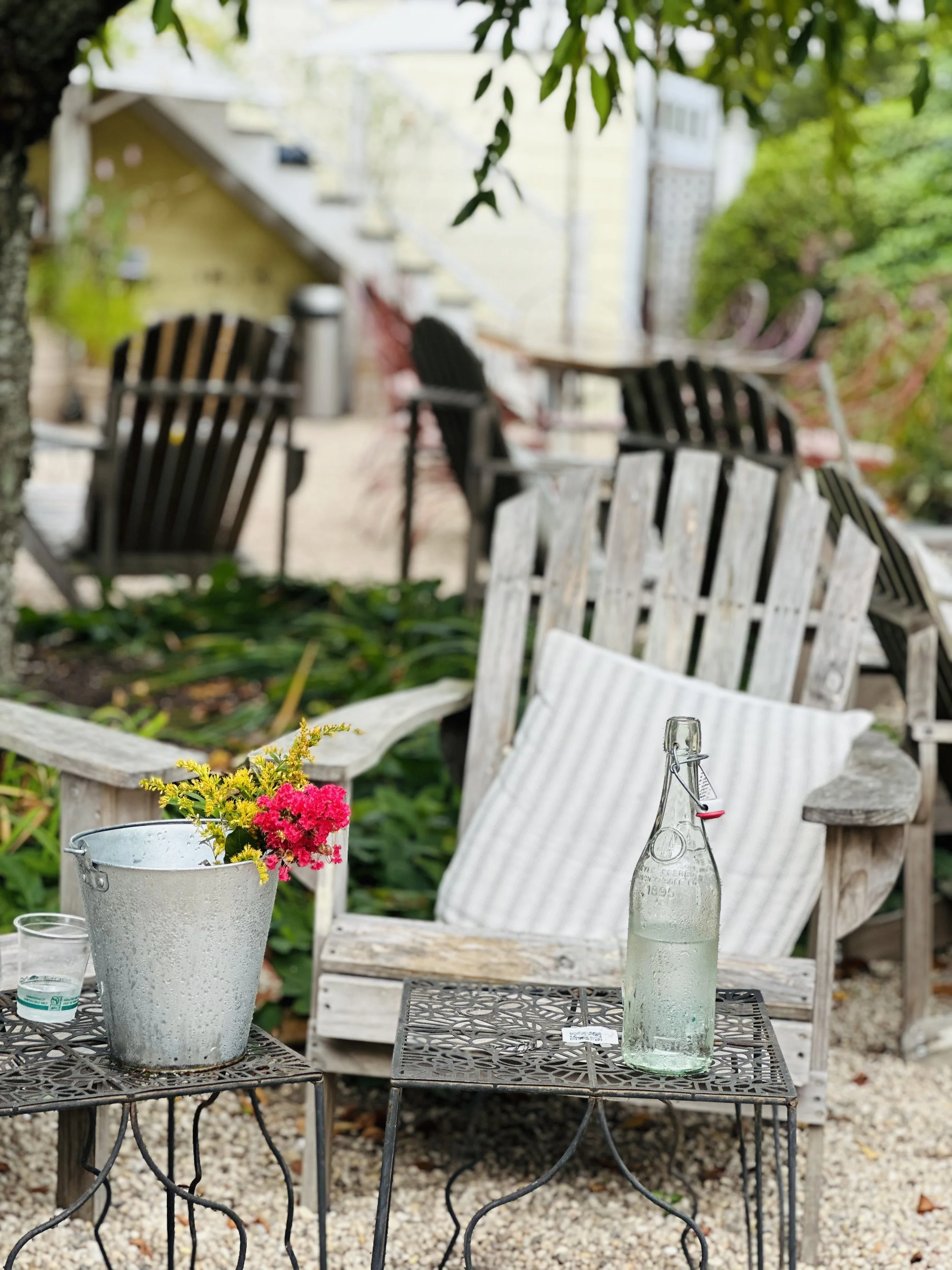 Outdoor patio scene with wooden chairs, a small table with a glass bottle of water, a striped pillow, a potted flower arrangement, and lush greenery in the background.
