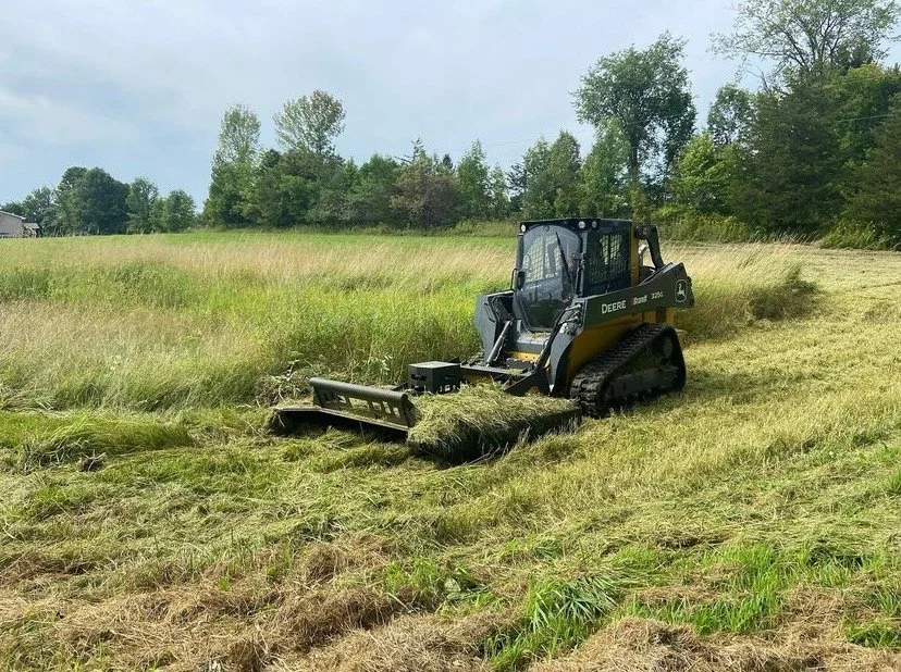 A small John Deere tracked skid steer loader mowing a field of grass.
