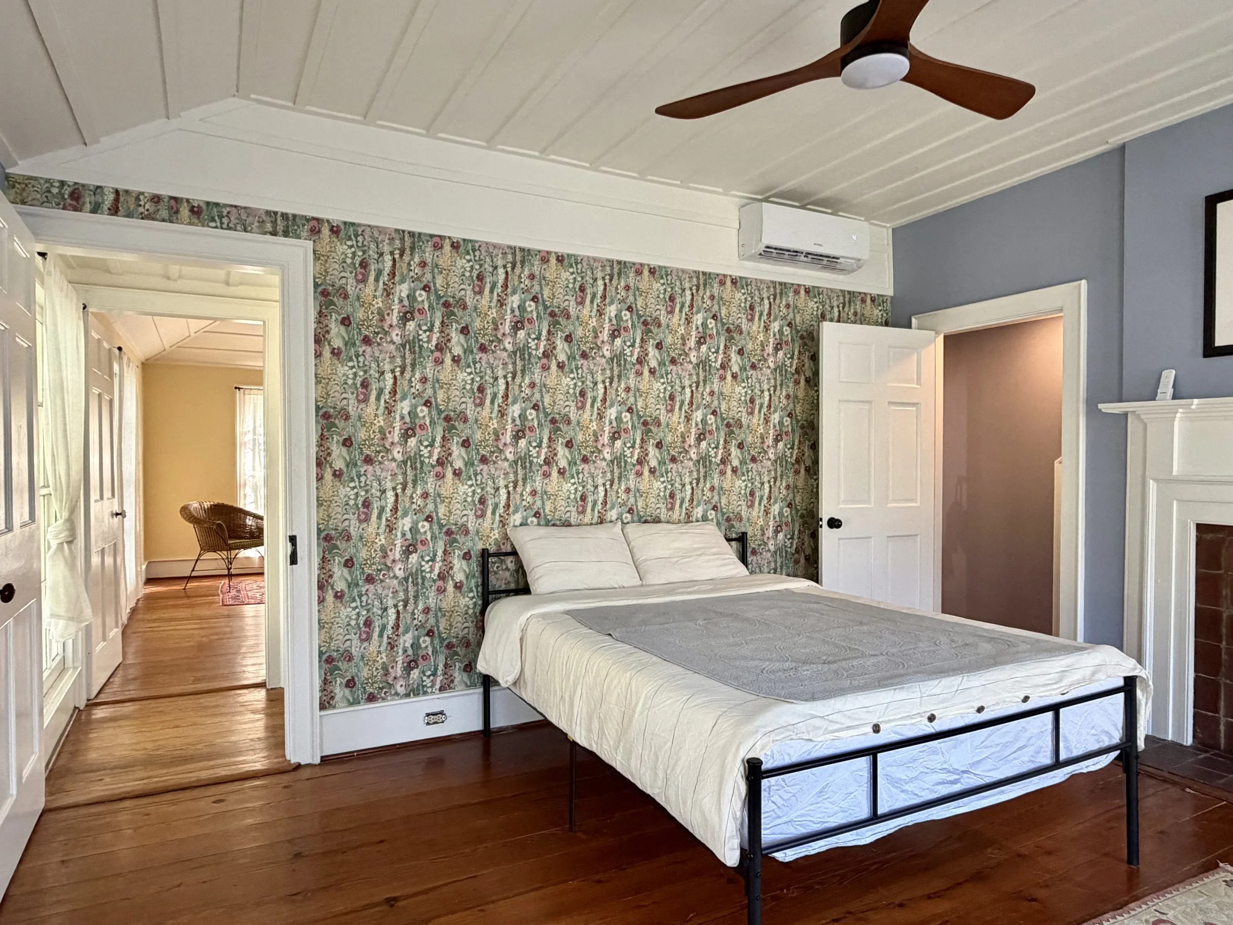 Upstairs bedroom with pale blue walls, floral wallpaper, a black metal bed, heart pine floors, and a white fireplace mantel.