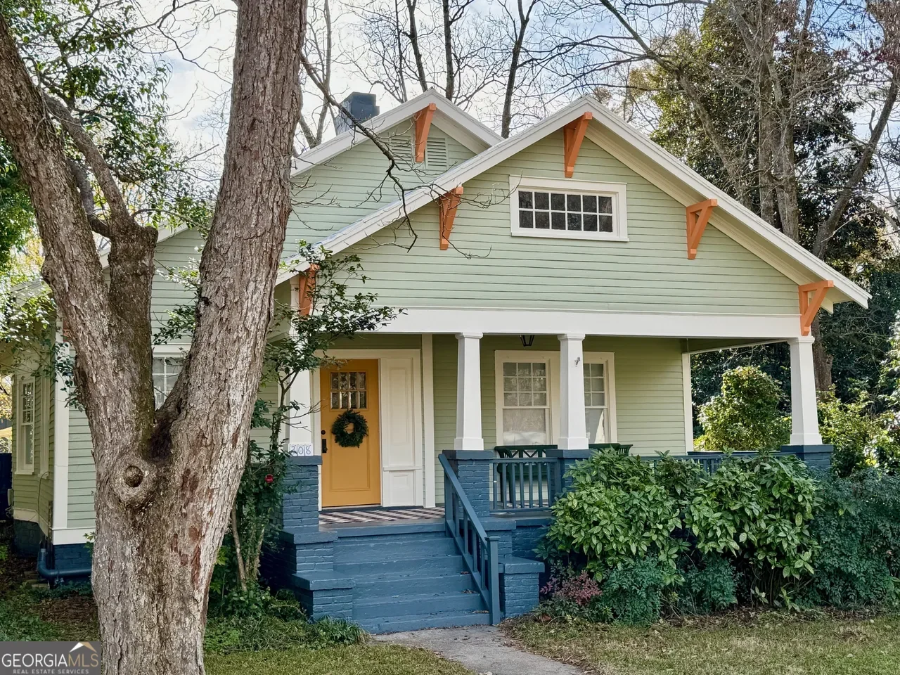 Historic pastel green cottage home in Washington, GA with wrap-around porch, orange door, blue steps, and large trees in a quiet neighborhood near downtown