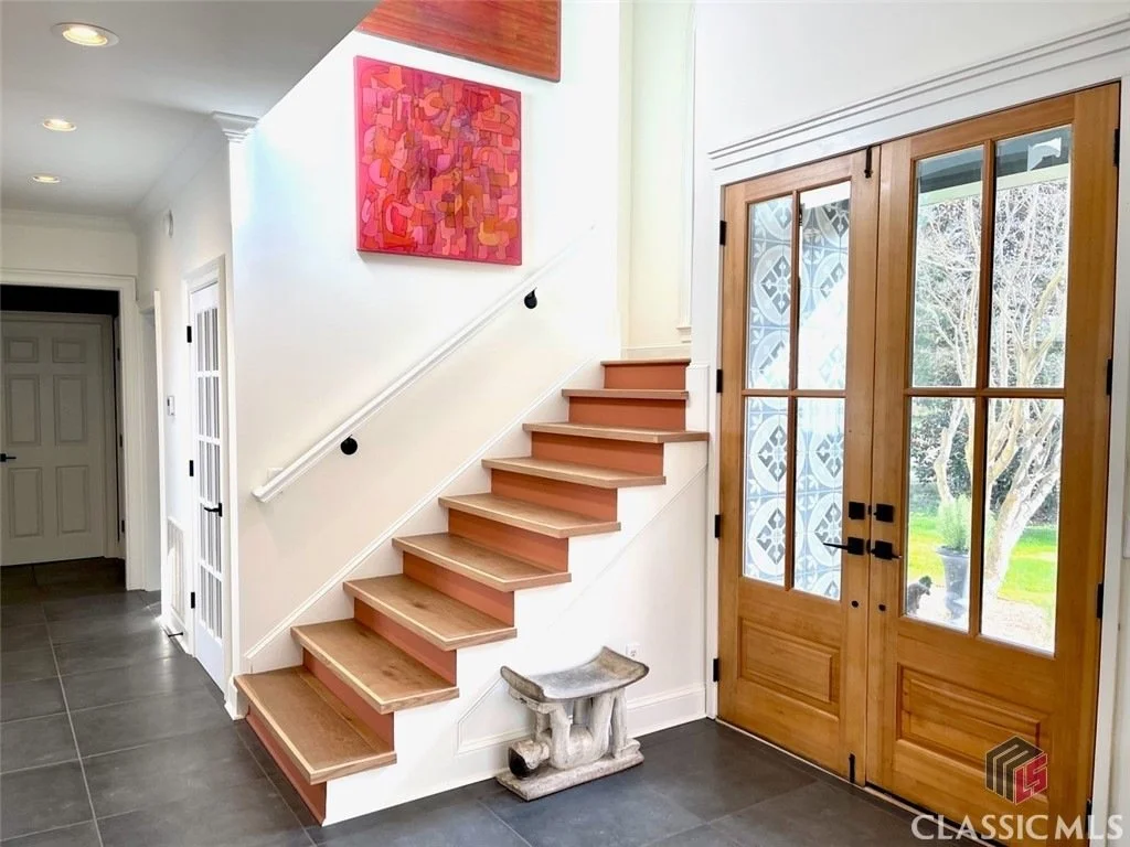 Interior view of a home's entryway featuring wooden double front doors with glass panels, a staircase with light wood steps and a white railing, a small stone sculpture underneath the staircase, a colorful abstract painting hung on the wall near the staircase, dark tiled flooring, and a glimpse of the garden outside through the glass