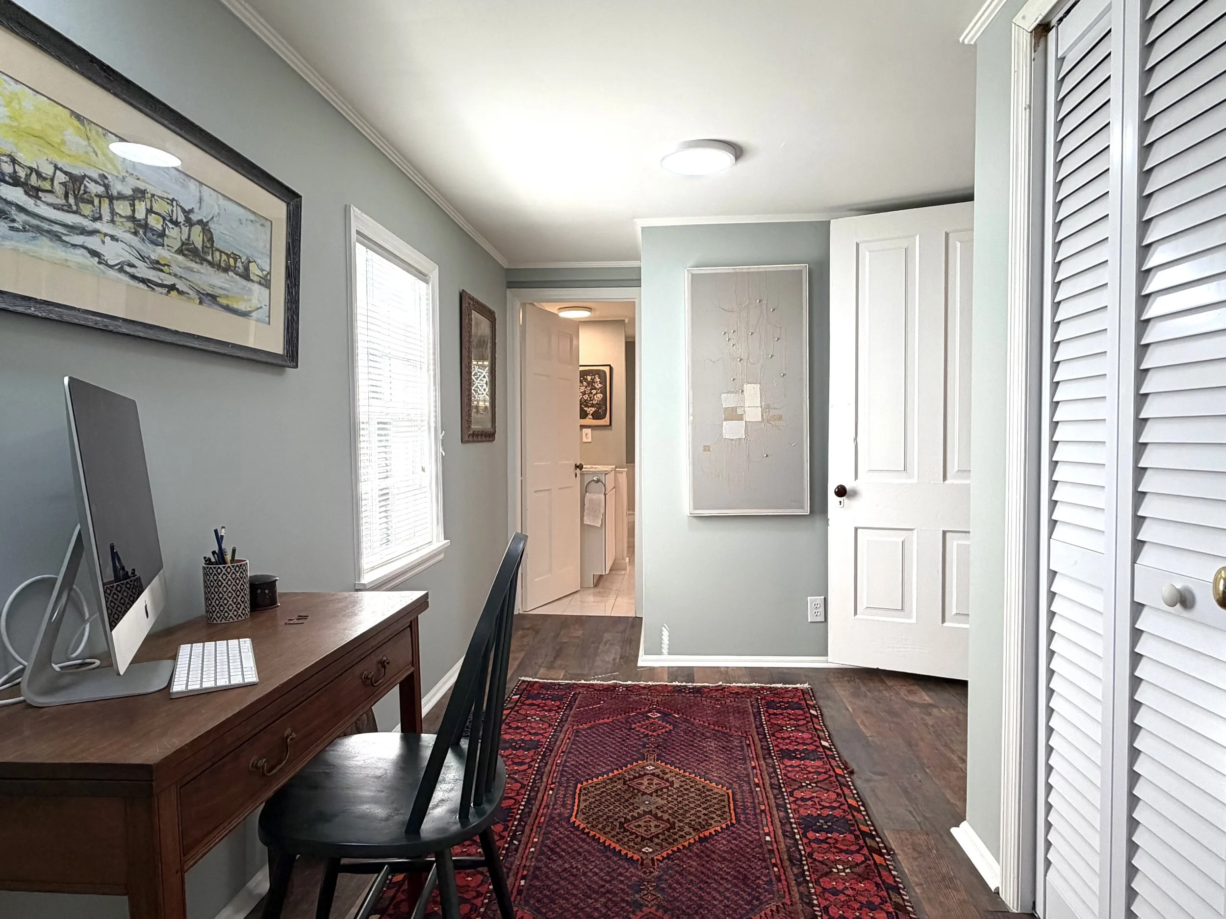 Home office with pale blue-gray walls, a wooden desk beside a window, white closet doors, and a red patterned rug.