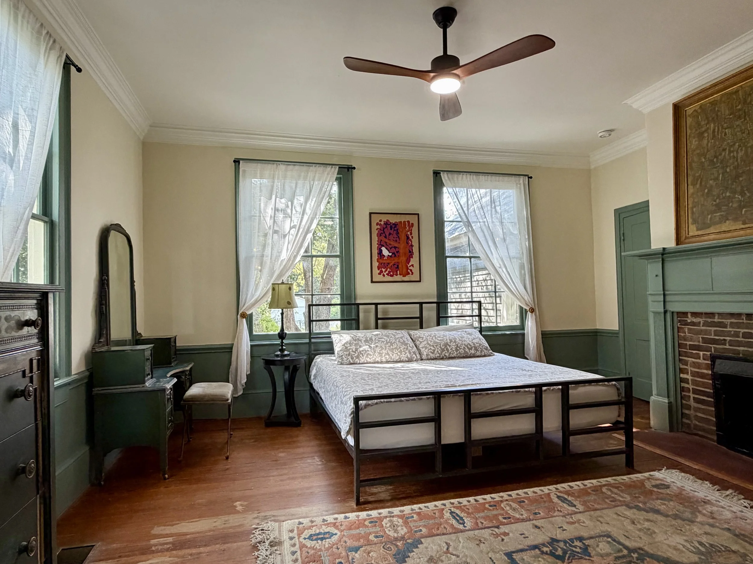 Primary bedroom with teal trim, tall windows, heart pine floors, and a decorative fireplace beside a black metal bed.