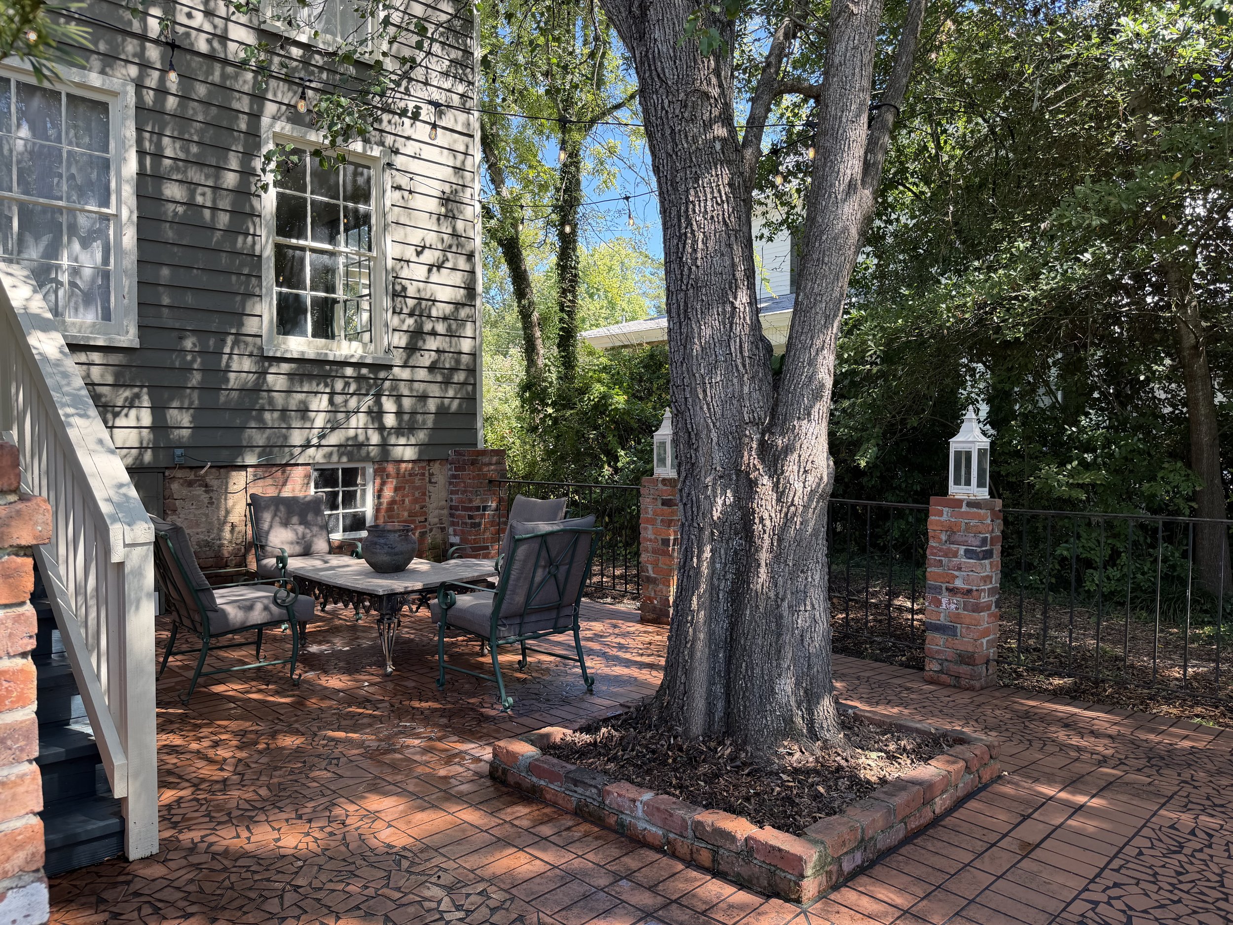 Brick courtyard with seating beneath a mature tree, overhead string lights, and iron railings between brick pillars.