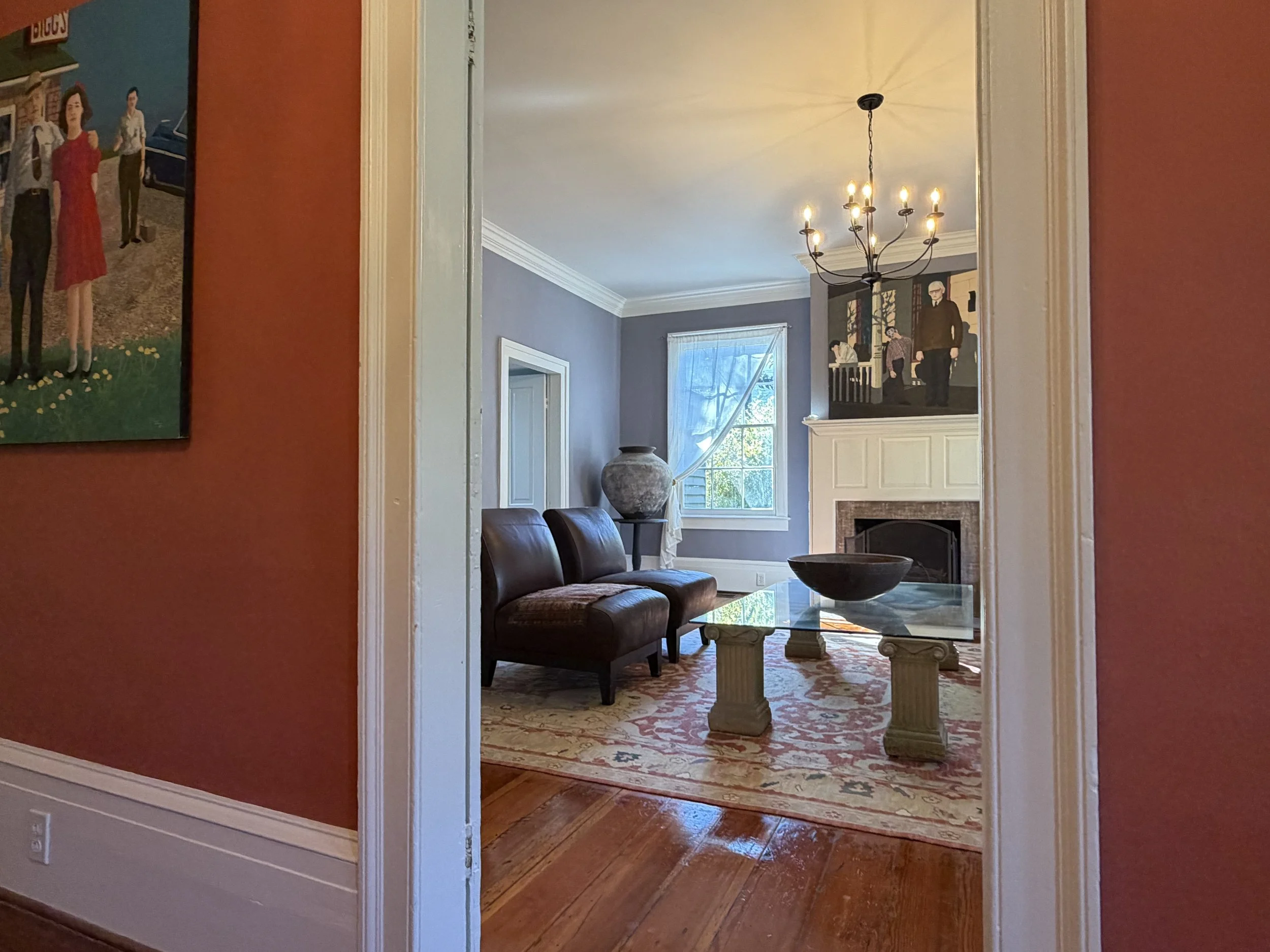 View through a white-trimmed doorway from the coral foyer into the living room, with a fireplace, chandelier, and leather chairs.