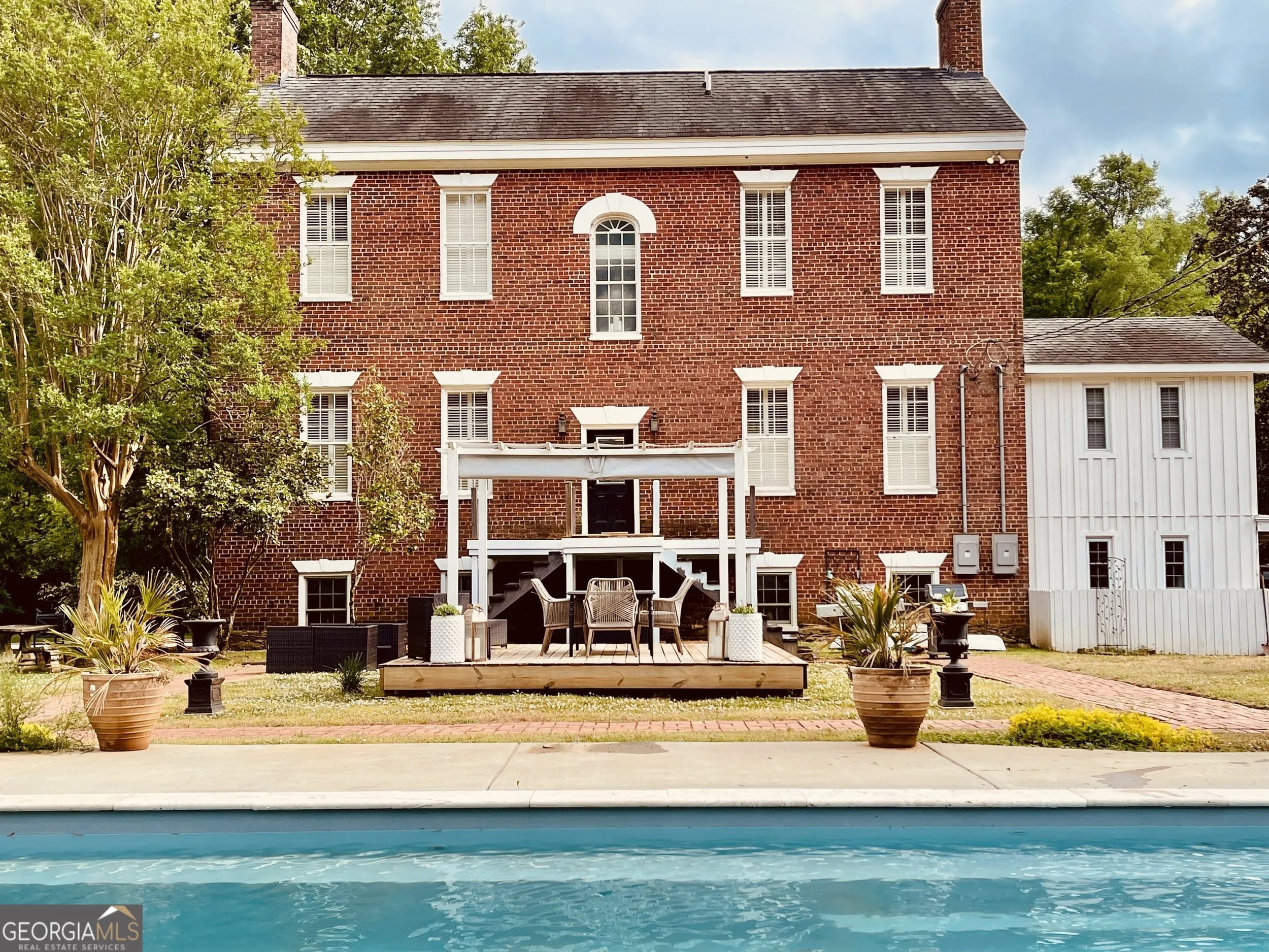The image shows a backyard with a brick house in the background, a wooden deck with outdoor furniture, potted plants, and a swimming pool in the foreground. Trees with green foliage are visible on both sides of the house.