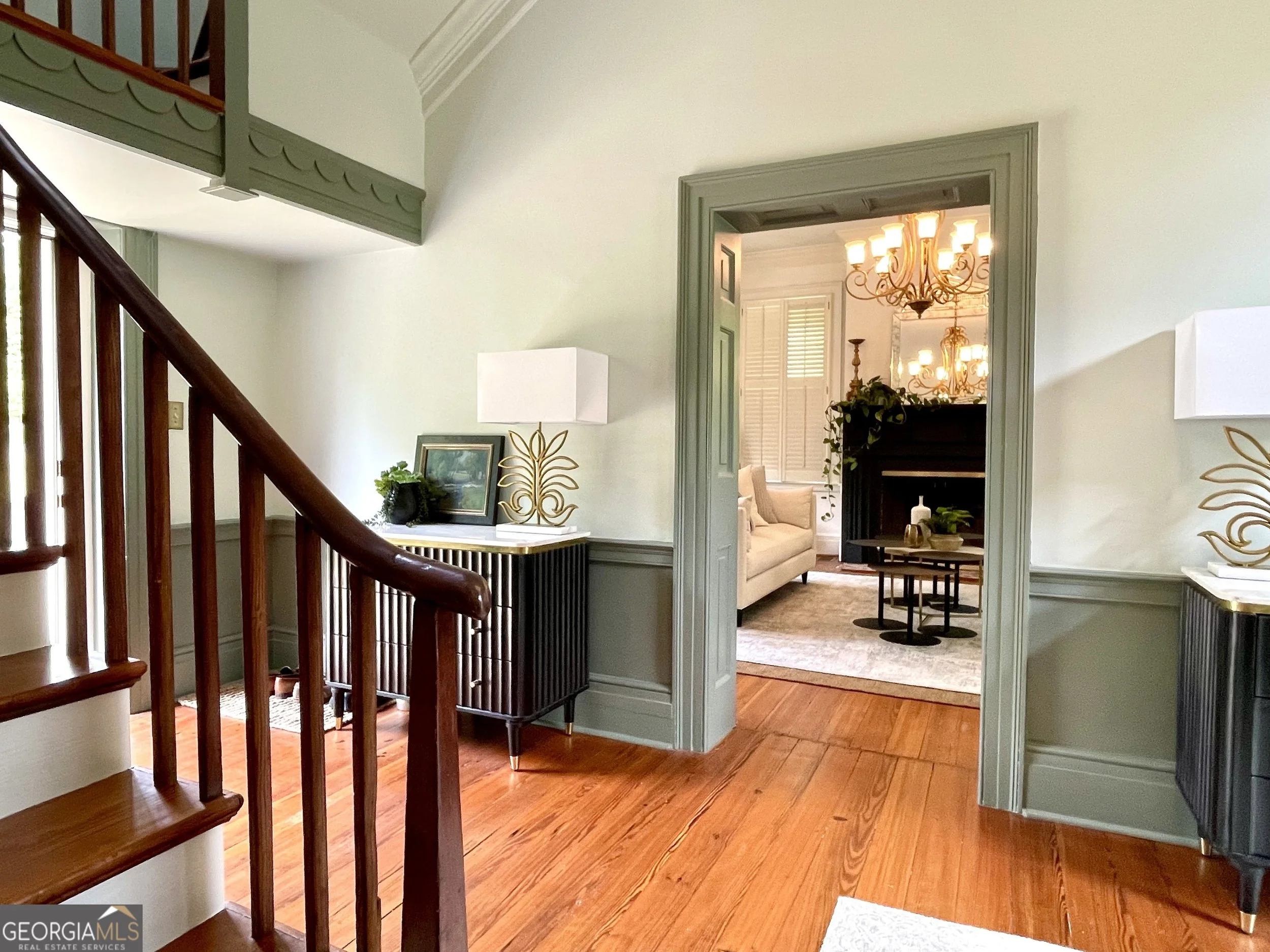 Interior view of a home with a staircase, a hallway, and a living room visible through an open doorway, featuring chandeliers, a white sofa, and a fireplace.