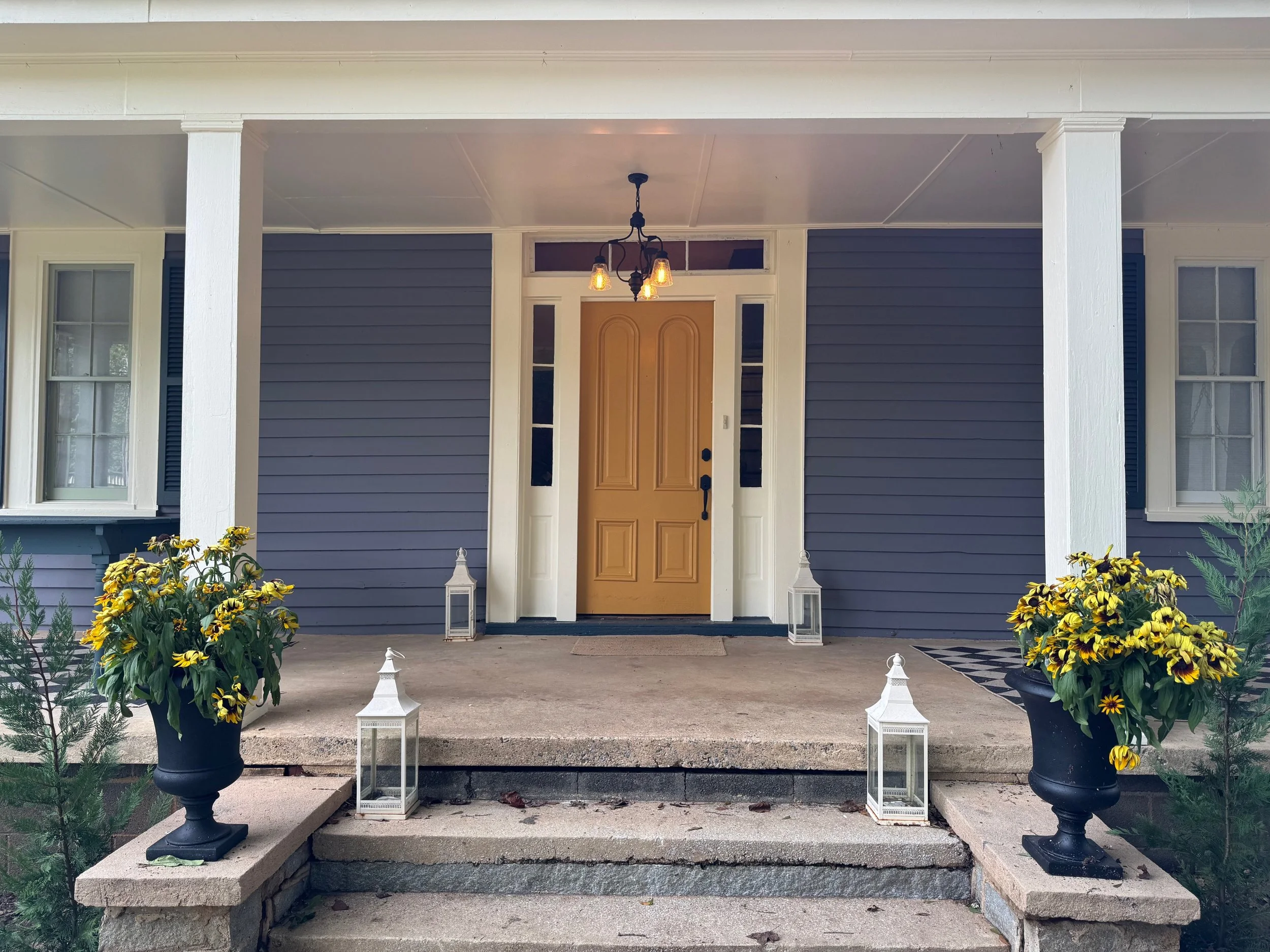 Front porch of a house with a yellow door, blue siding, white trim, hanging light fixture, and decorated with yellow flowers and white lanterns.
