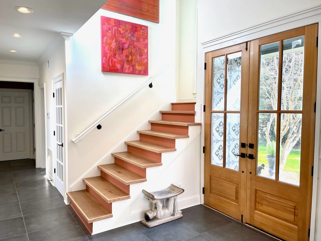 Interior entryway with a wooden front door with glass panels, a staircase with wooden steps and a white railing, a colorful abstract painting on the wall, and a small stone bench near the door.