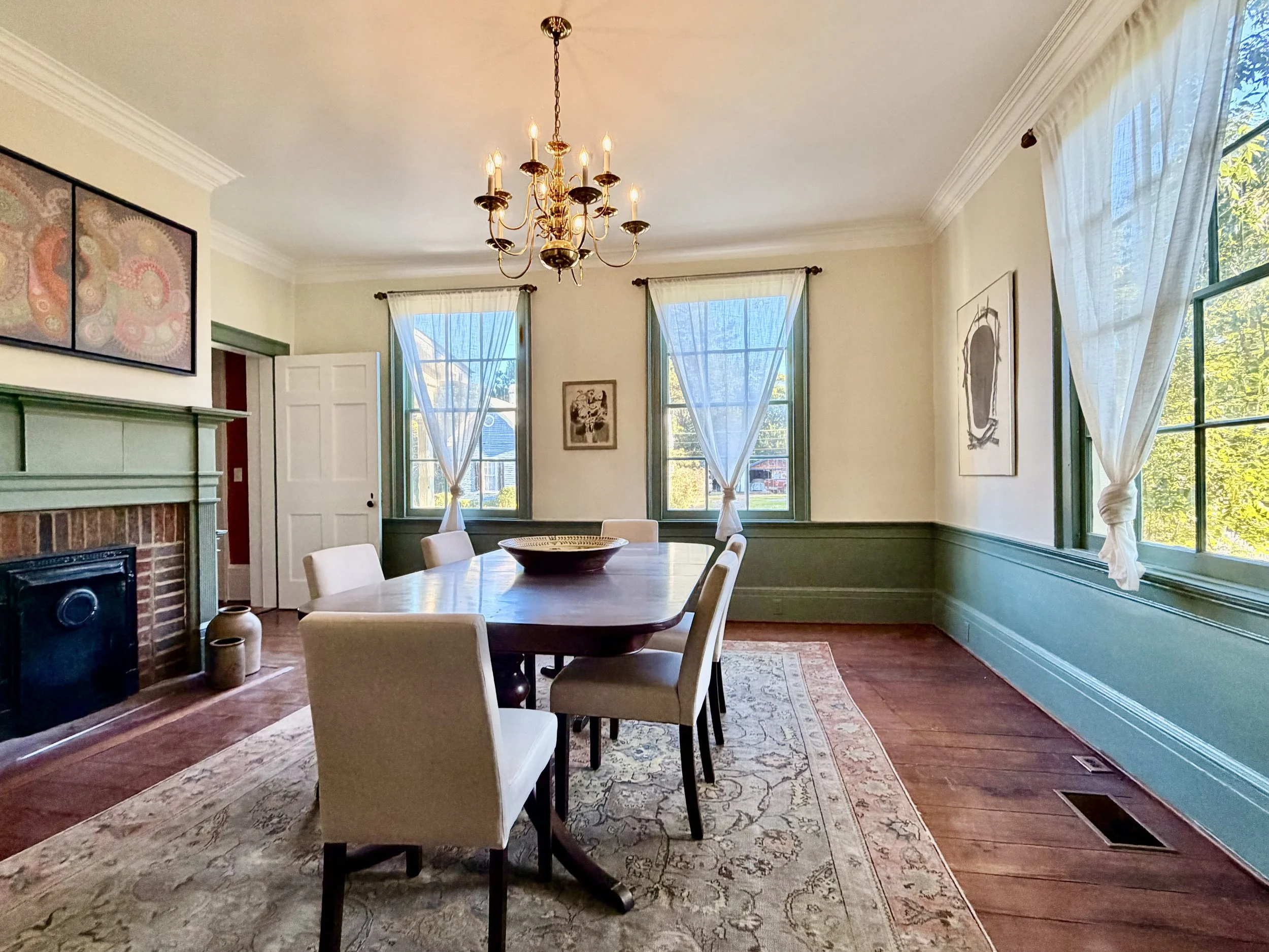 Sunlit dining room with a decorative fireplace, teal wainscoting, tall windows, and a brass chandelier above the table.