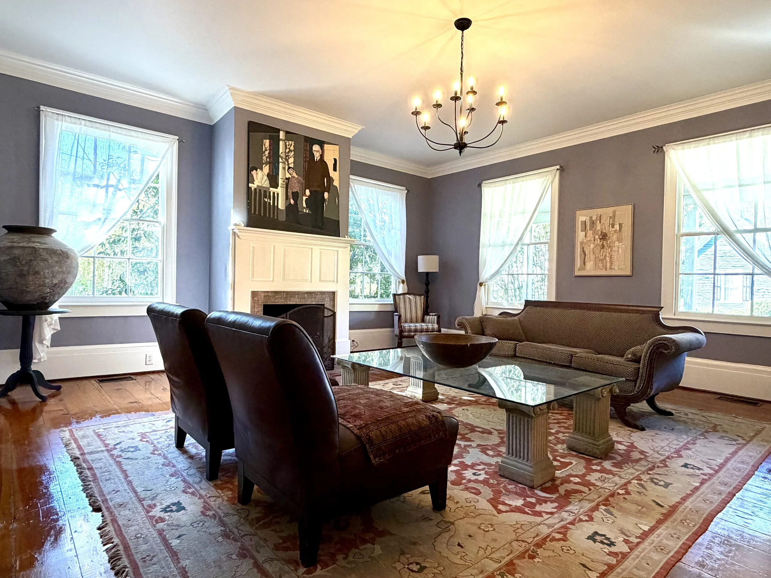 Living room with lavender walls, a white fireplace mantel, leather chairs, and an ornate sofa beside tall windows.