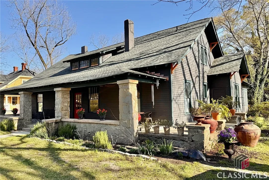 Charming historic Craftsman-style cottage home exterior in Lexington, Georgia featuring gray siding, white trim, wrap-around porch with stone pillars, red front door, green porch roof, large trees, and lush landscaping in a peaceful neighborhood