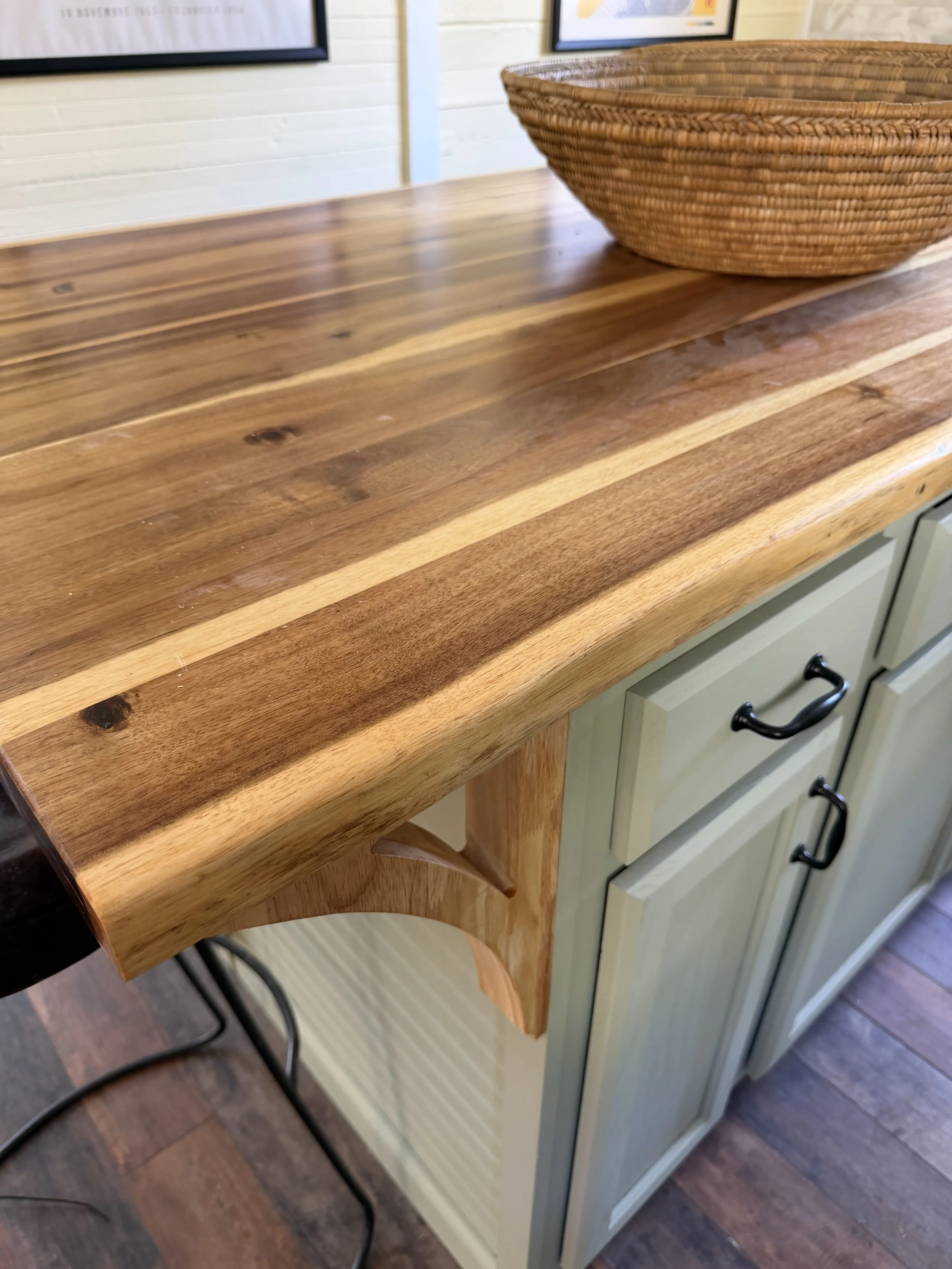 Close-up of the live-edge wood countertop above pale green kitchen cabinetry, with a wooden support bracket and woven basket.