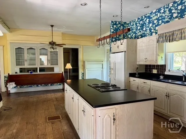 Kitchen with white cabinets, black countertop island, refrigerator, window with floral valance, and hanging pot rack. Yellow walls and wooden floors.