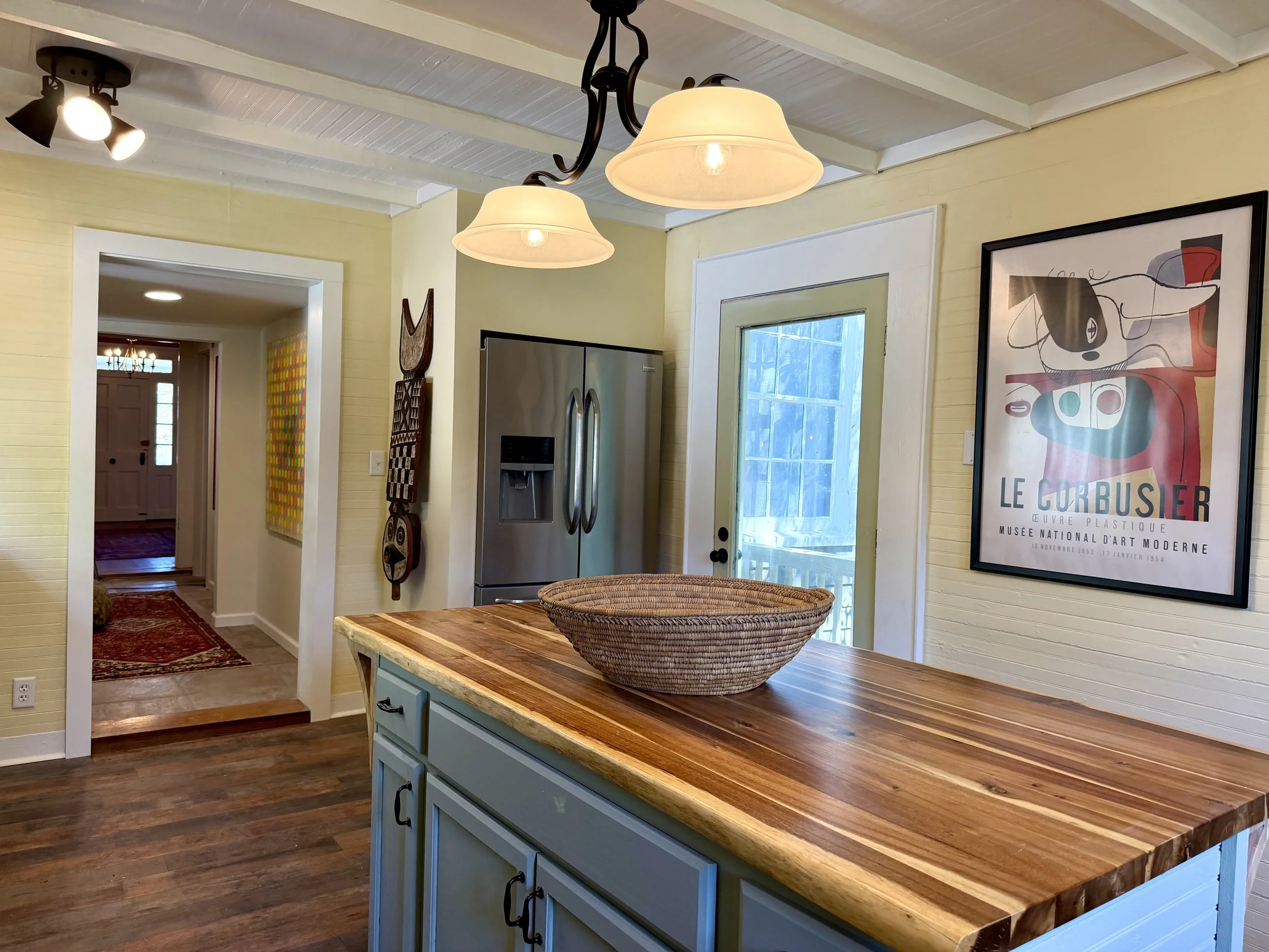Live-edge wood countertop on a pale green kitchen island beneath pendant lights, with framed artwork and a view toward the foyer.