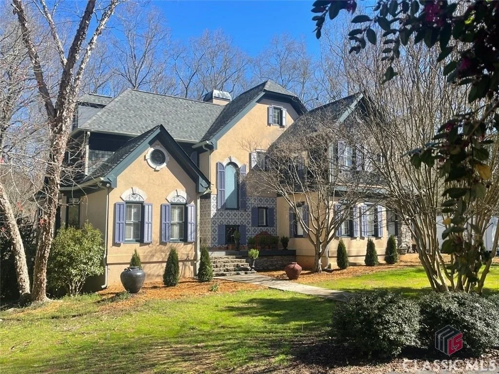 A large, elegant house with a gabled roof and multiple windows with blue shutters, surrounded by leafless trees, shrubs, and potted plants, under a clear blue sky.