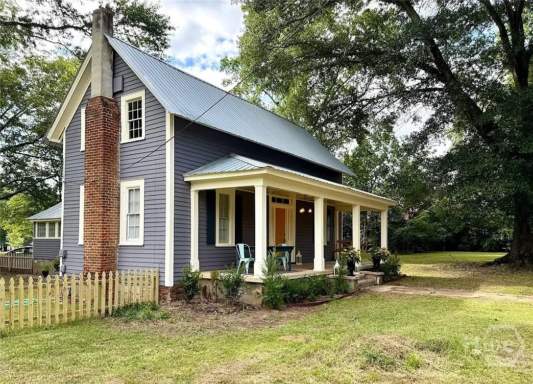Charming historic cottage-style home exterior in Crawford, Georgia featuring gray siding, white trim, wrap-around front porch with columns, brick chimney, tin roof, picket fence, and mature oak trees