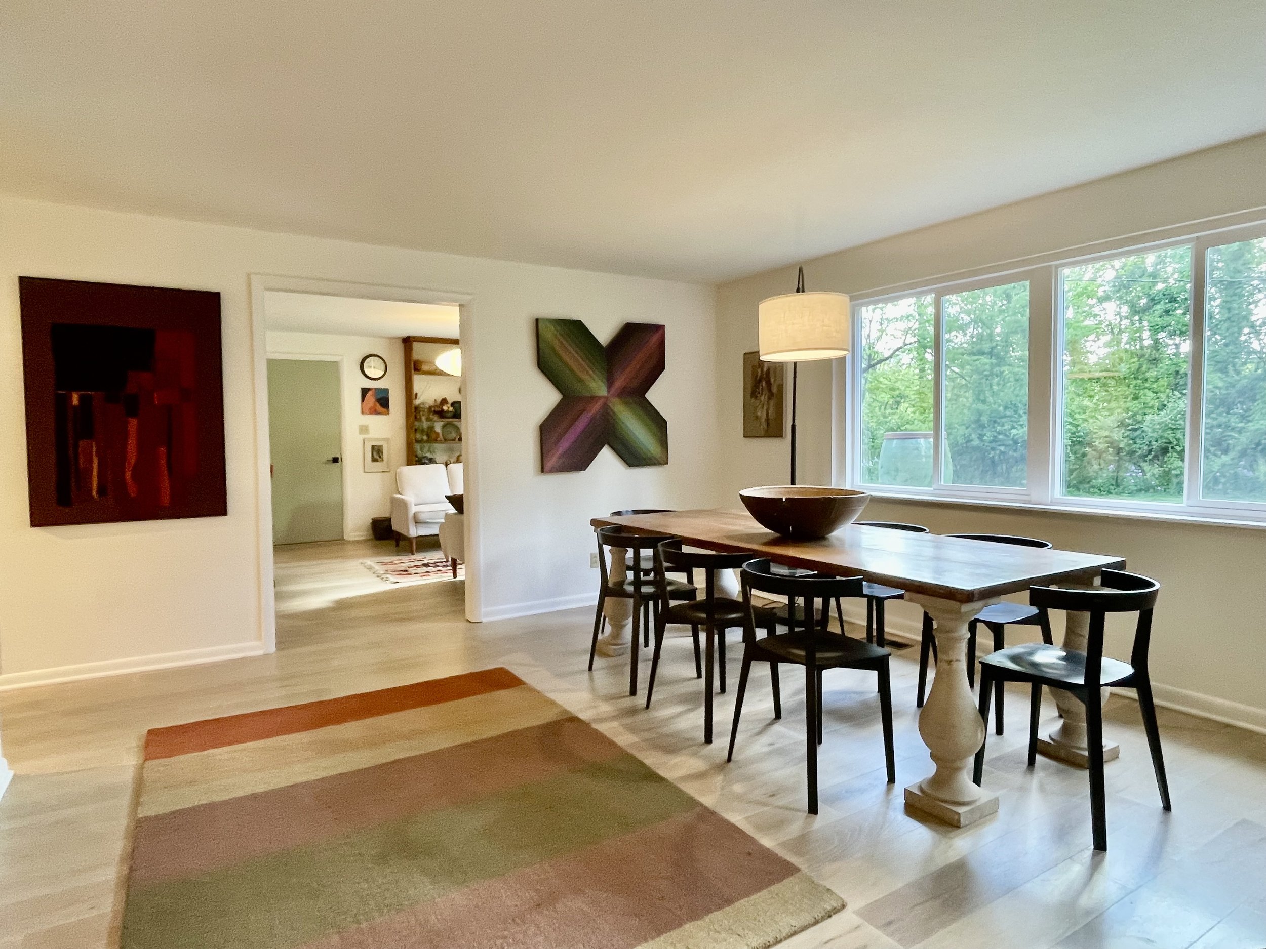 A dining room with a wooden table and black chairs, large window with greenery outside, colorful abstract wall art, white walls, and a light-colored ceiling.