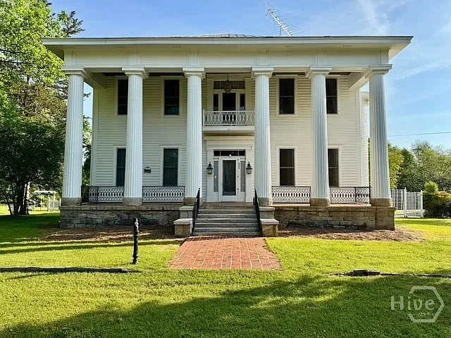 A large white Southern-style mansion with tall columns at the front entrance, brick steps leading up to it, surrounded by a green lawn and trees under a blue sky.