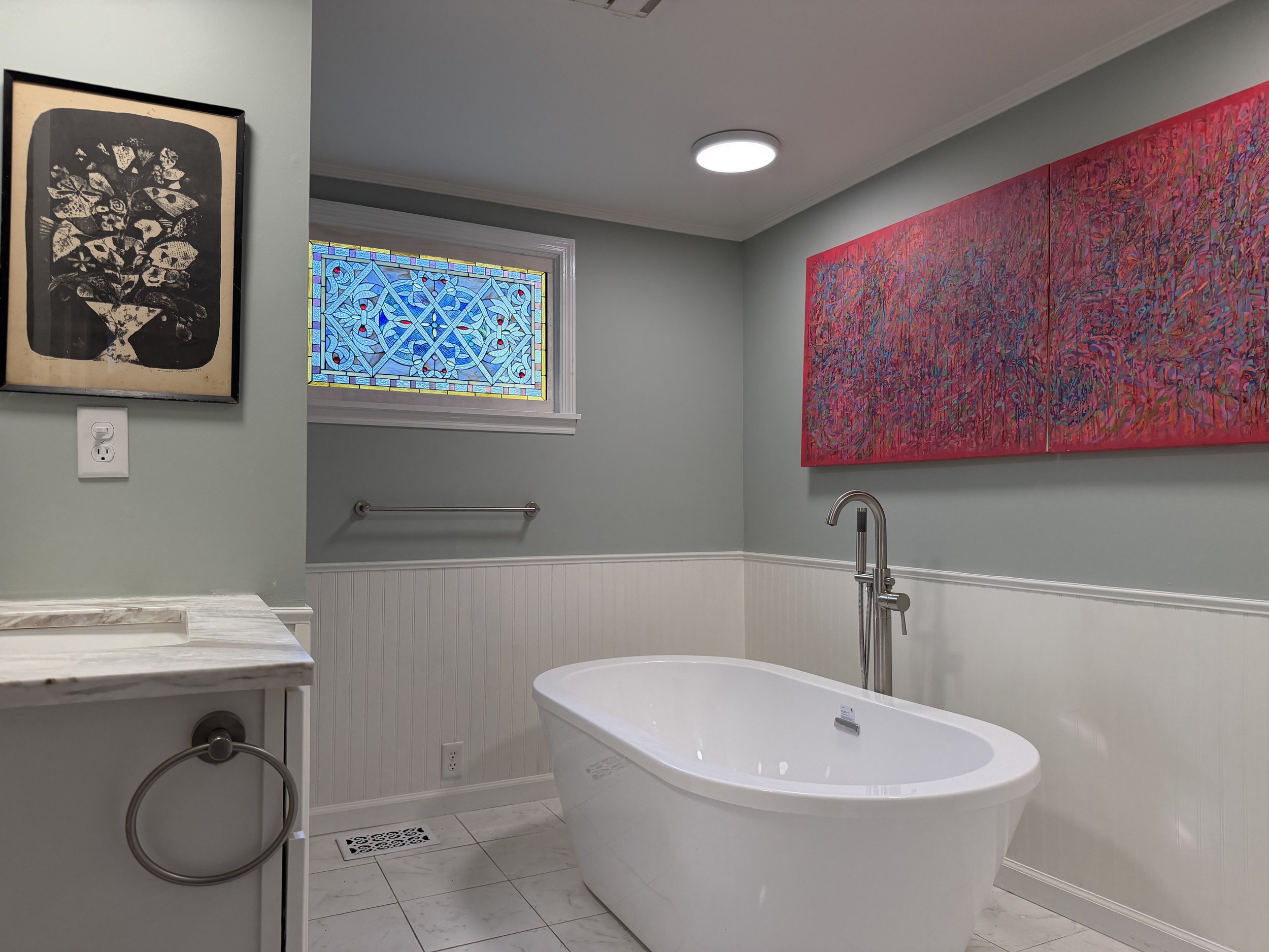 Freestanding white soaking tub beside a blue stained-glass window, with white wainscoting and colorful wall art.