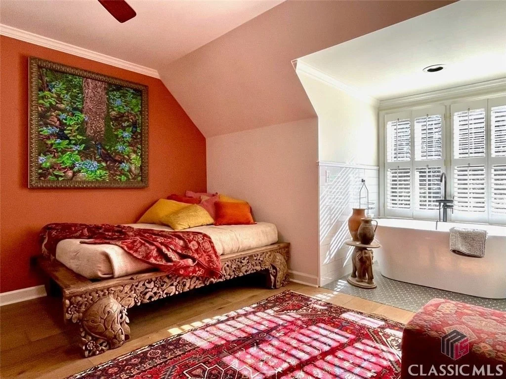 Decorative interior room with a daybed against an orange wall, colorful pillows, a framed nature-themed artwork, a patterned rug, a white tub in a windowed alcove with shutters, and vintage-style vases.