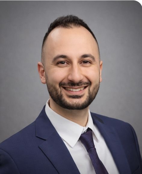 Professional man in a blue suit, white shirt, and purple tie smiling against a gray background.