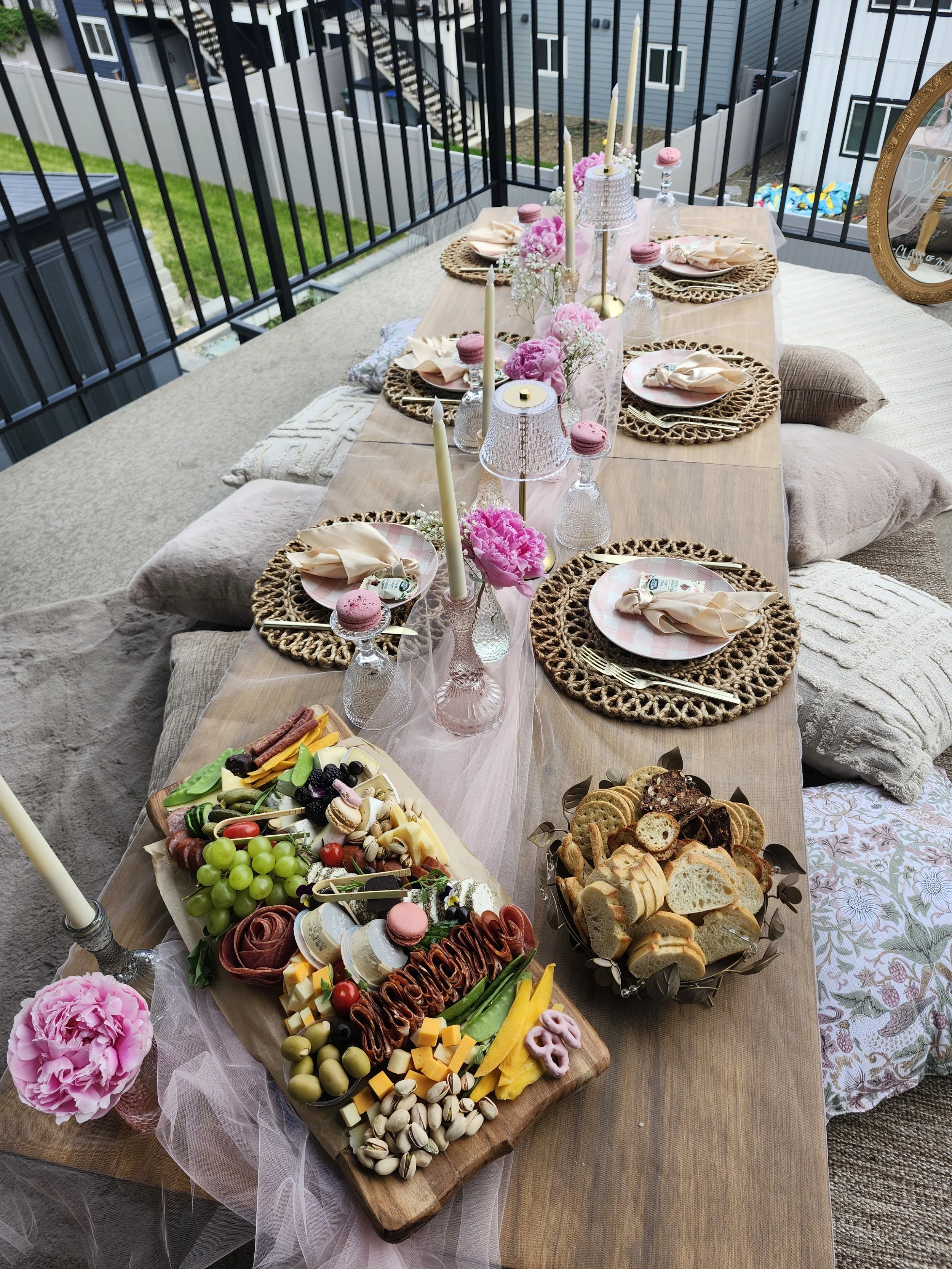 A backyard outdoor dining table set for a celebration, with pink flowers, candles, and a cheese and fruit platter, surrounded by cushions and a blanket.