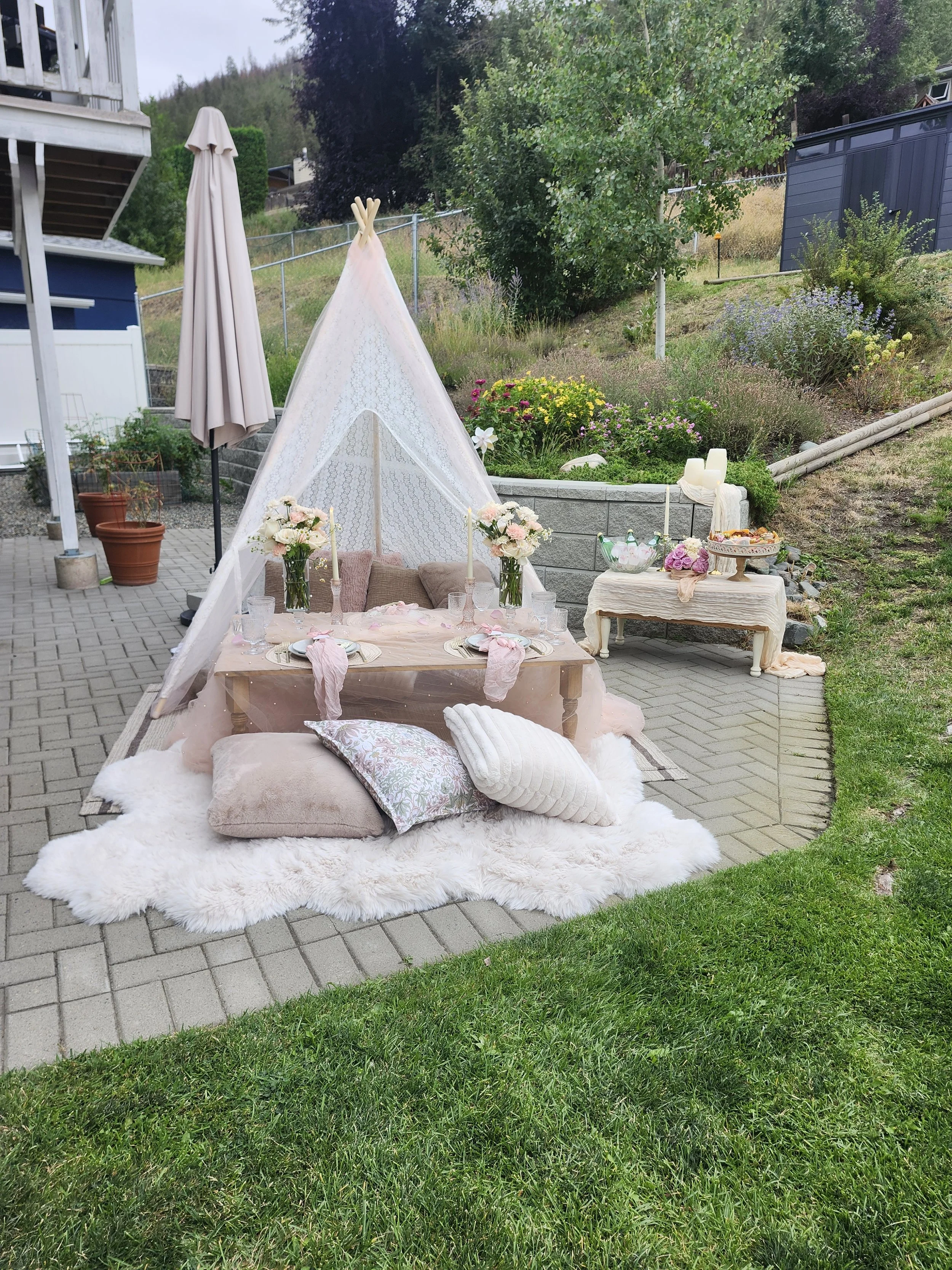 A backyard scene with a table set for a small gathering, decorated with flowers and candles, placed on a white fluffy rug on paving stones, surrounded by grass and trees, with a teepee tent in the background.