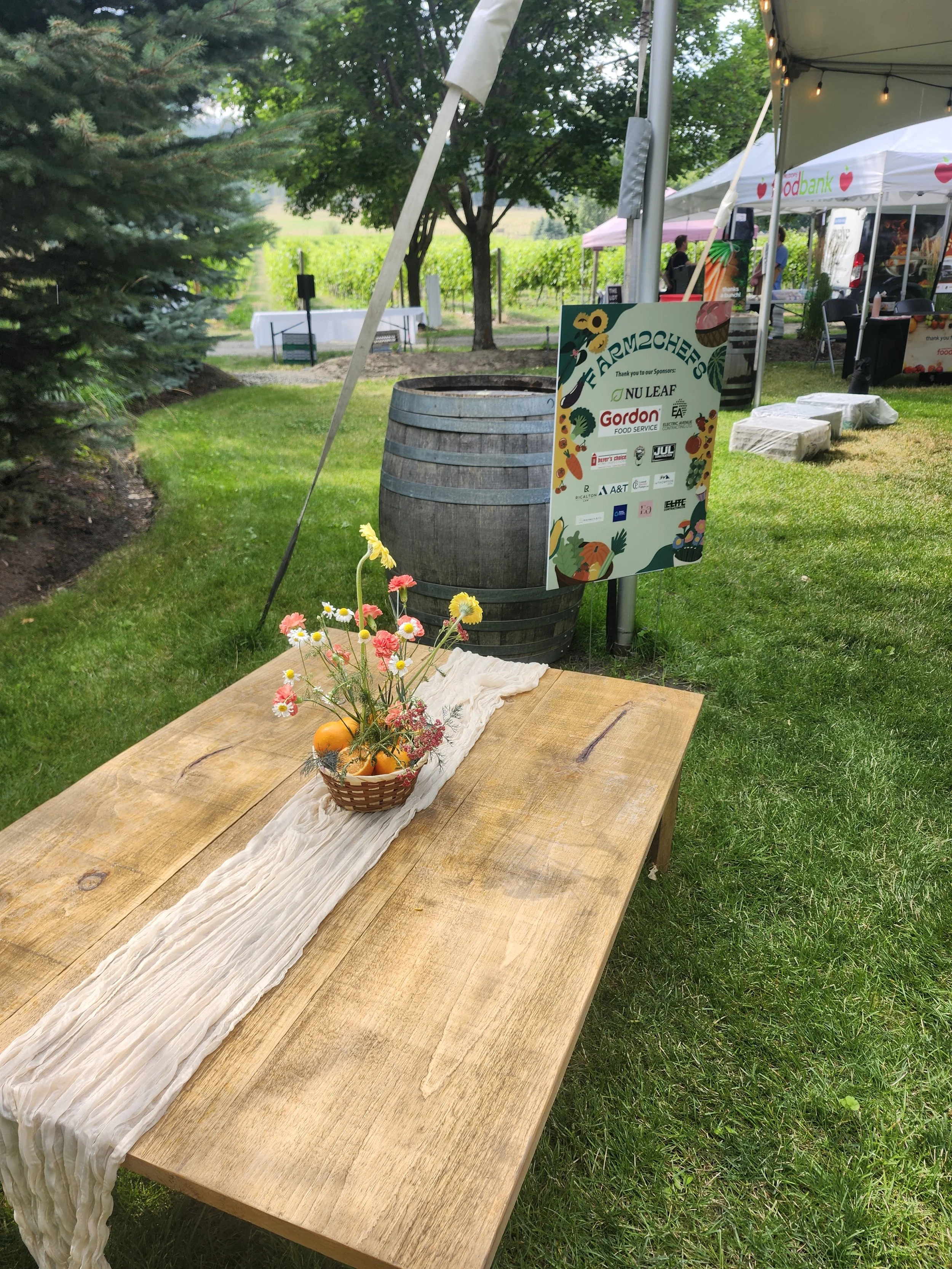 A wooden table with a cream-colored cloth runner and a small basket of flowers and pumpkins, set outdoors at a farmer's market, with tents and greenery in the background.