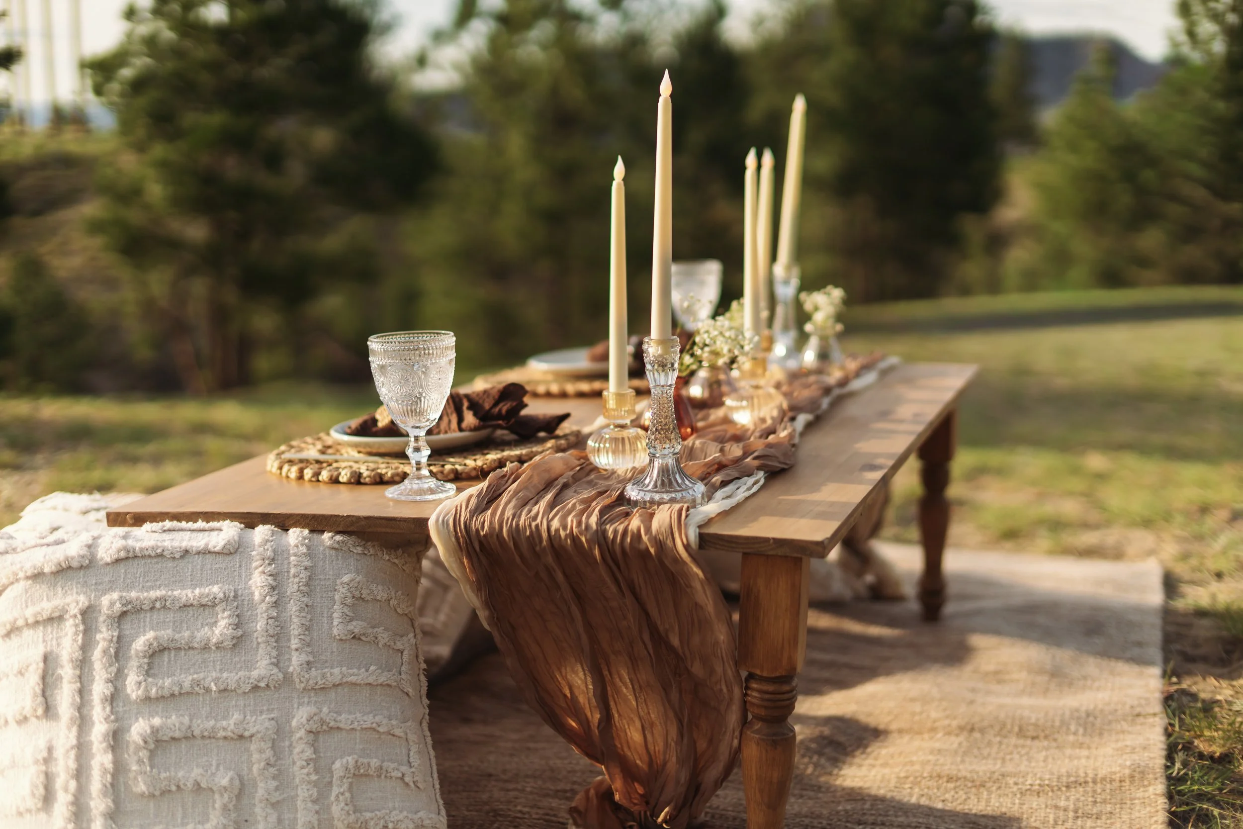 Outdoor table set with white candles, glassware, flowers, and a textured table runner on a wooden table, with a green natural background.