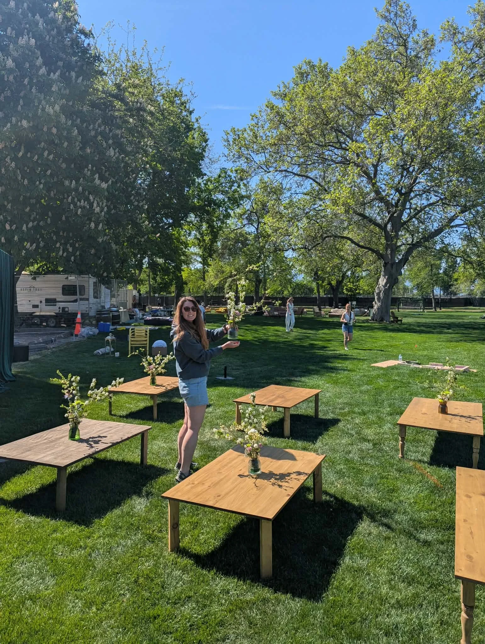 A woman holding a glass jar with flowers at an outdoor event set up with low wooden tables and vases of flowers, across a grassy field with large trees and a clear blue sky.