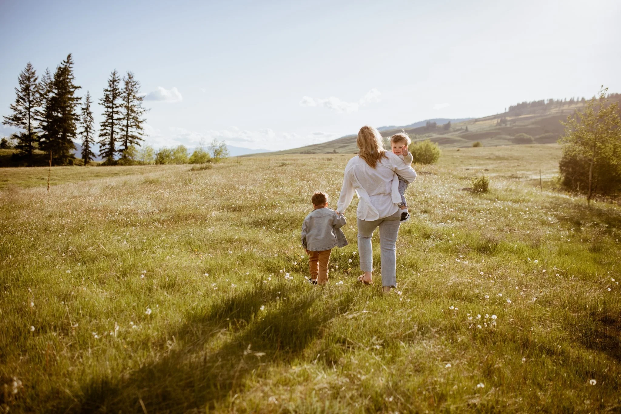 A woman walking in a grassy field with two children on a sunny day, one child holding her hand and the other riding on her back, with trees and hills in the background.