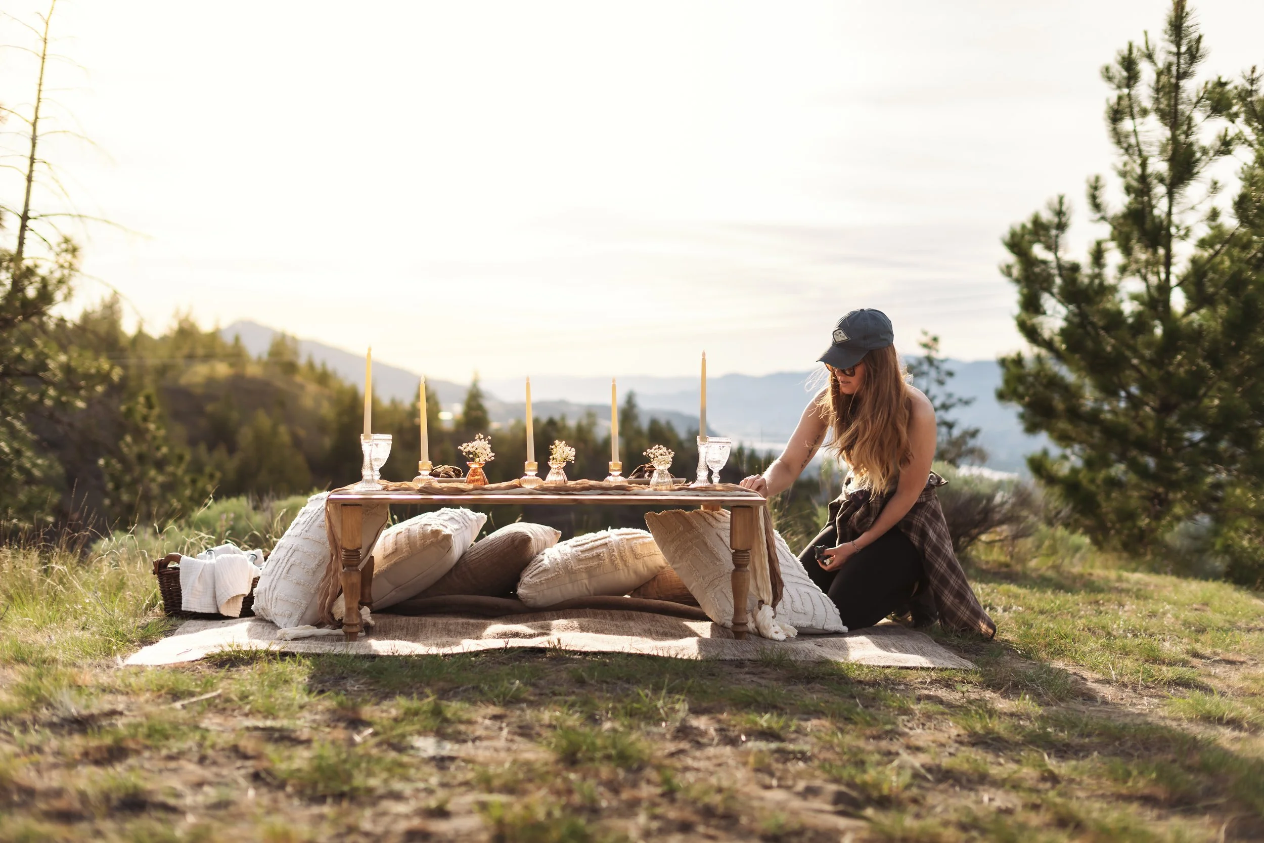 A woman with long hair wearing a baseball cap and glasses is sitting and kneeling beside a low wooden table set outdoors in a grassy field with trees and mountains in the background, decorated with candles and flowers for a rustic outdoor gathering.