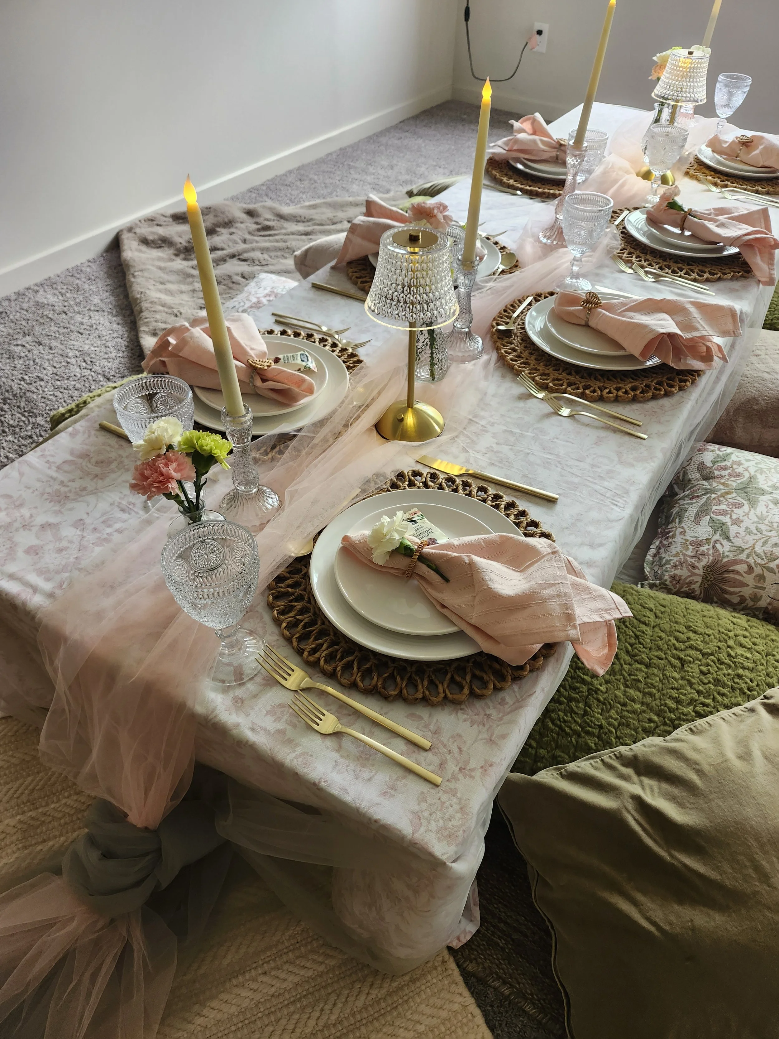 A decorated dining table with pink napkins, white plates, gold flatware, glassware, candles, and flowers, set for a celebration or dinner.