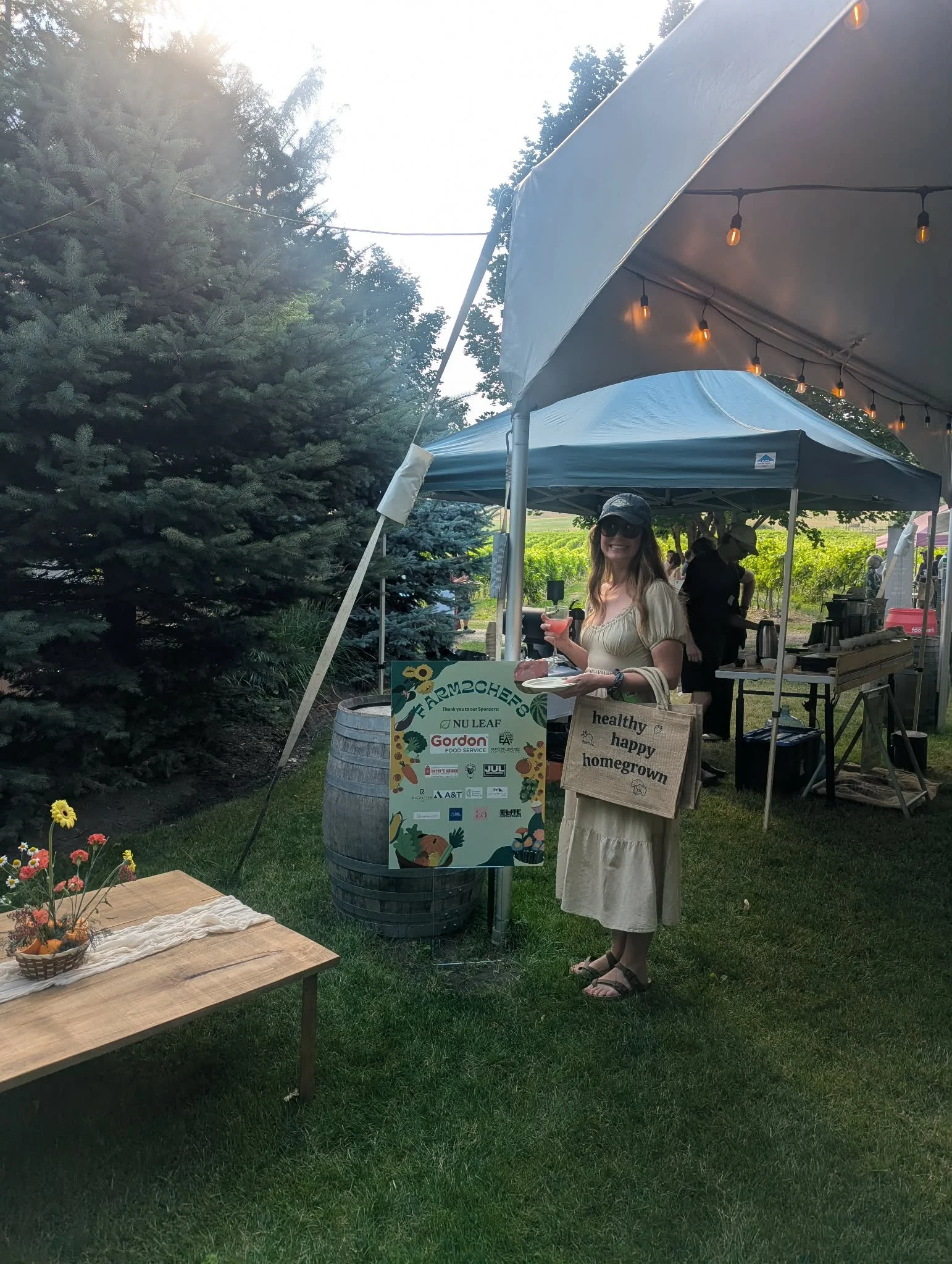 A woman wearing sunglasses and a hat, holding a pink drink, standing outdoors at a market or festival under a canopy, with a display sign that reads 'Farmers'. There is a table with a basket of flowers and a cloth in the foreground, trees, and other 