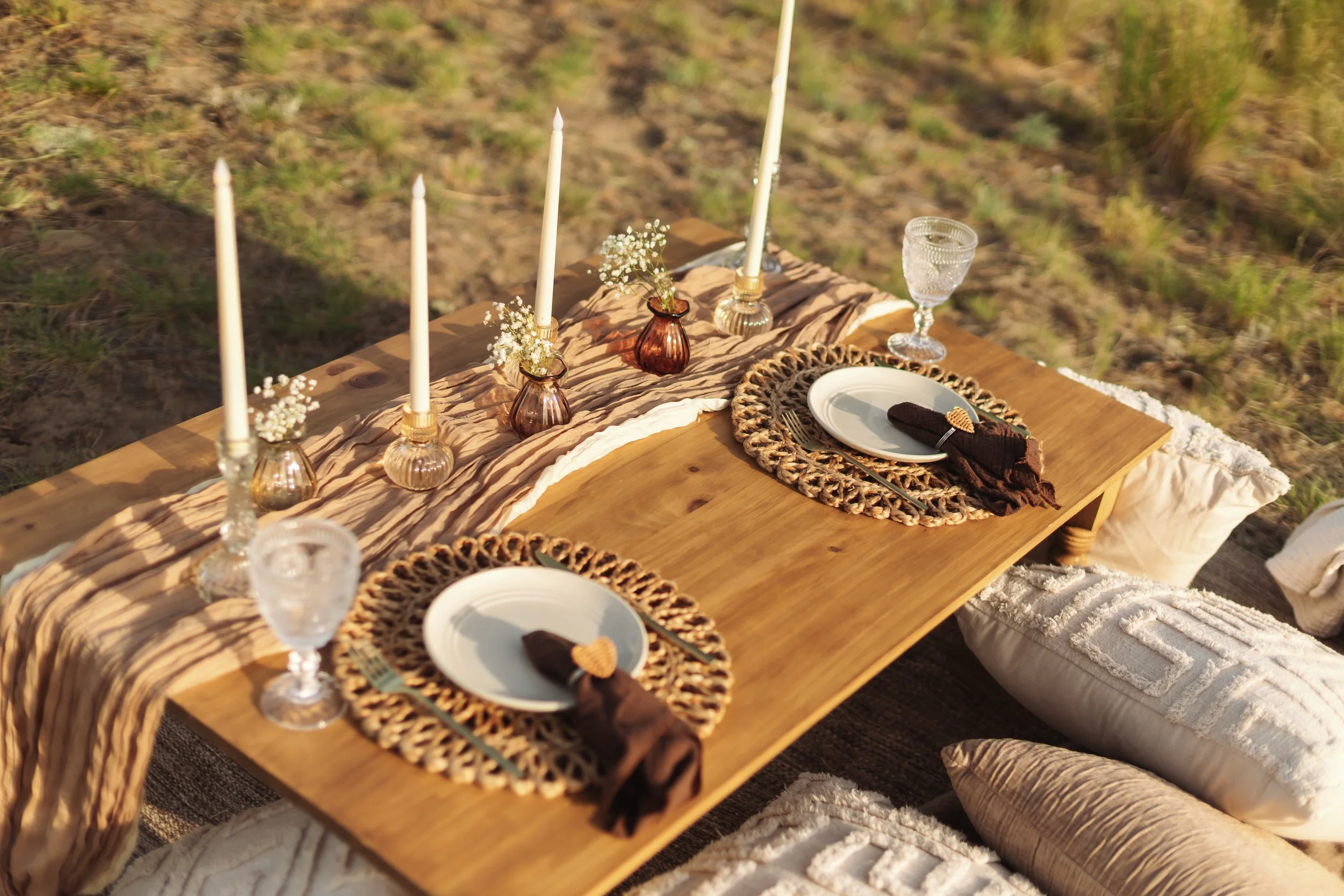 An outdoor dining setup on a wooden table with candles, small flower arrangements in brown vases, white plates with brown napkins, and glassware, on a sandy surface with grass in the background.