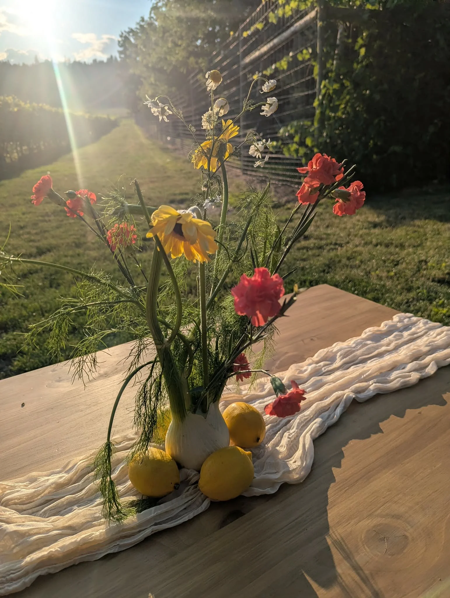 A bouquet of wildflowers and lemons in a white vase on a draped cloth, set on a wooden table outdoors with sunlight and green garden in the background.