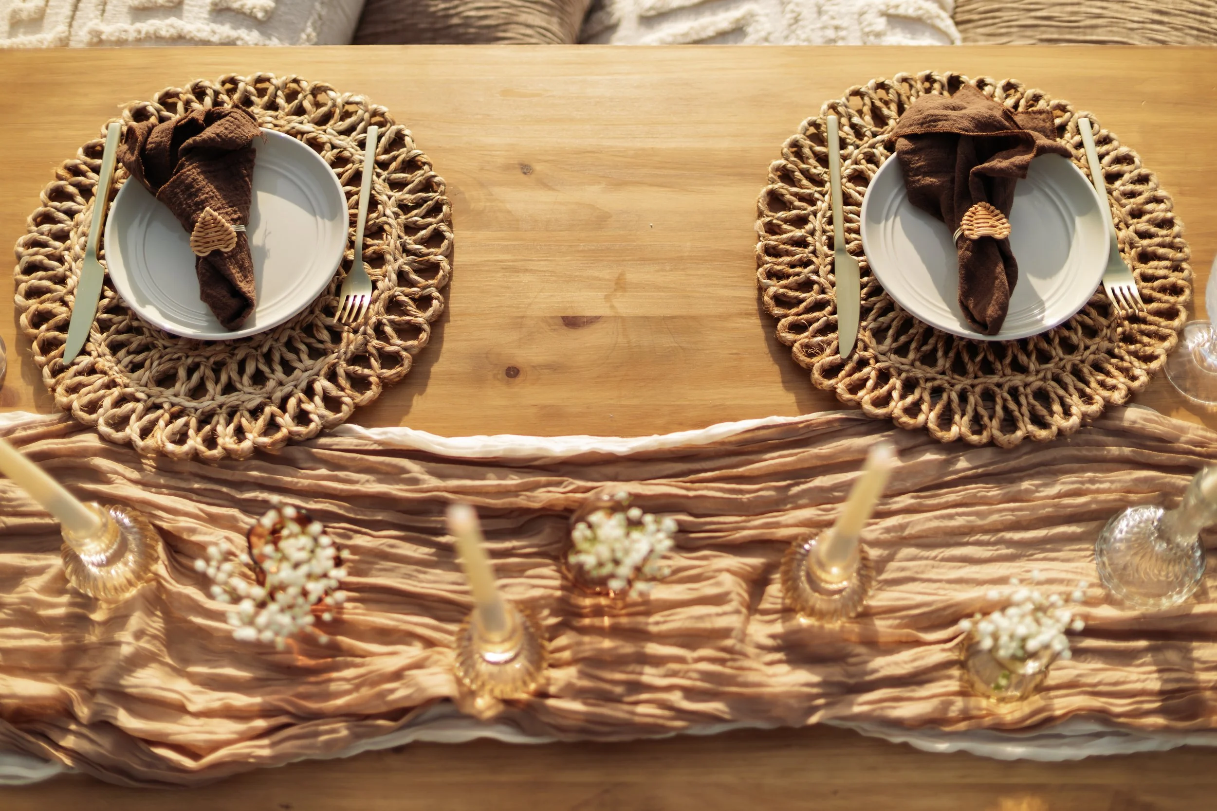 Two place settings on a wooden table with woven placemats, white plates, brown cloth napkins, and gold utensils, arranged for a meal.