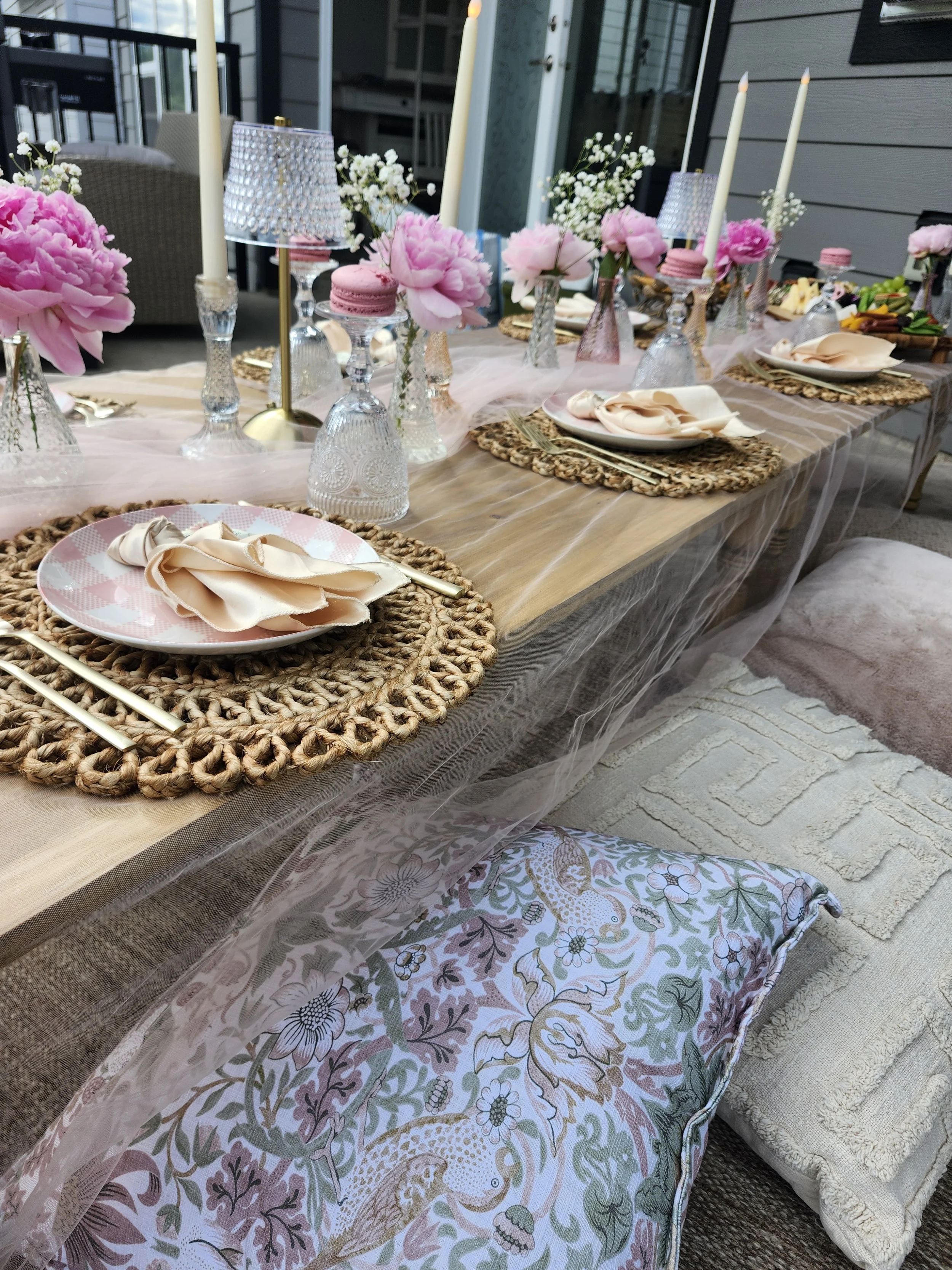 A decorated outdoor dining table with pink peony flowers, white candles, pink macarons, and various candelabras, set on woven placemats, with patterned pillows and a textured rug underneath.
