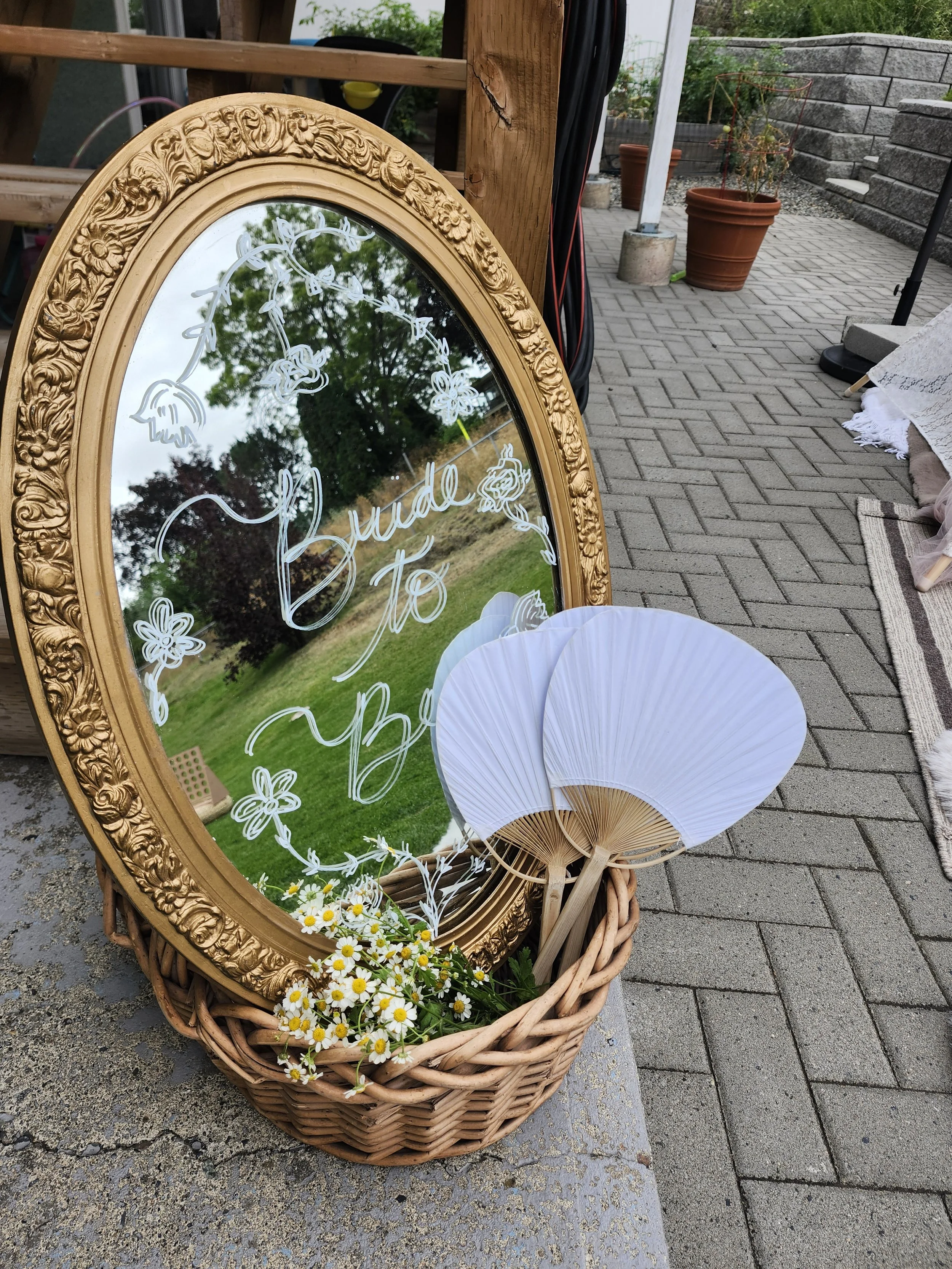 A decorative gold-framed mirror with white floral designs and the words 'Bride To Be' written on it, set on a wicker basket filled with white daisies with yellow centers. Two white paper parasols are leaning against the basket, and the scene is outdo