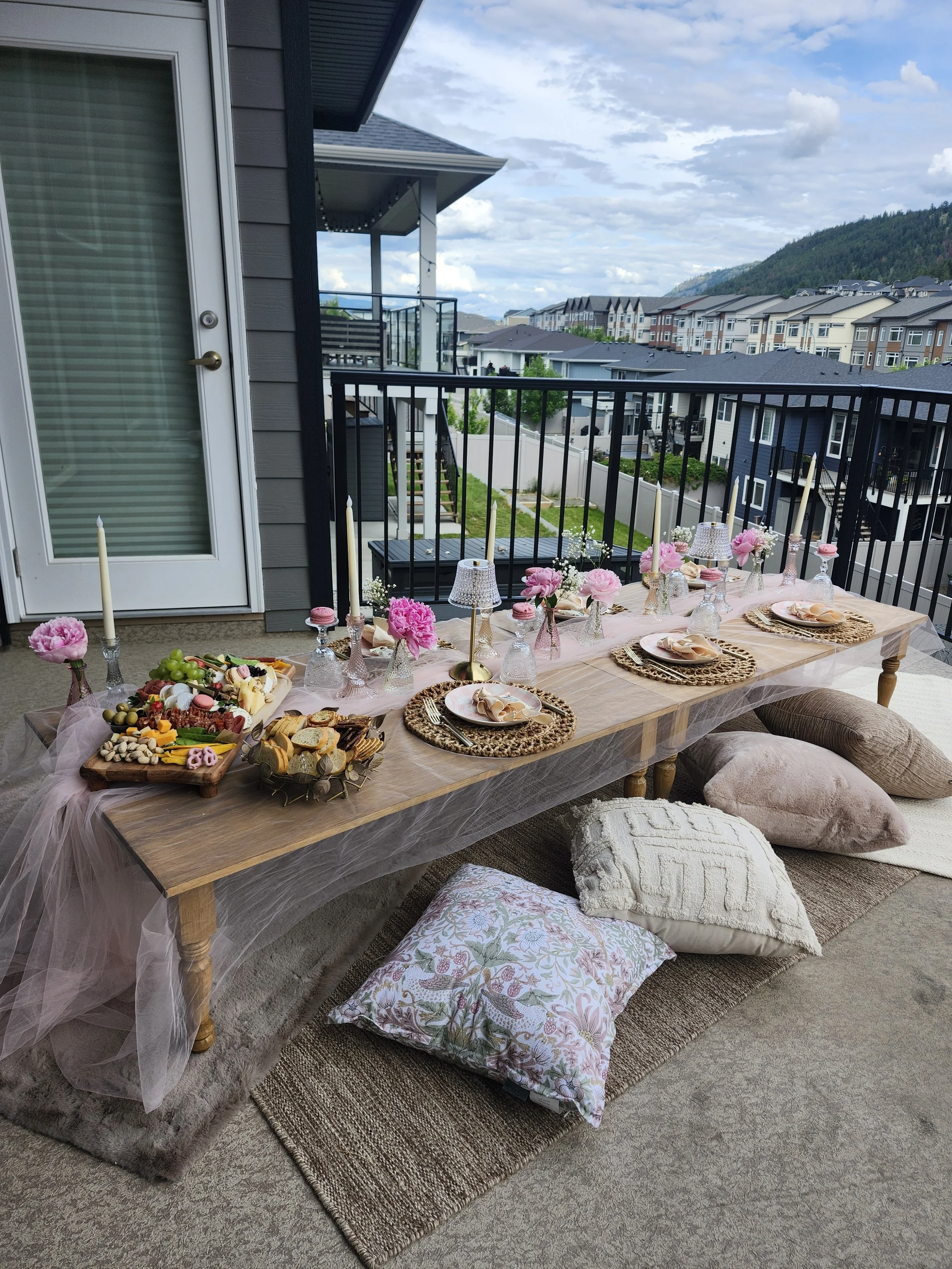 An outdoor patio with a wooden table decorated for a celebration, featuring pink flowers, candles, and refreshments. The table is covered with a pink tablecloth and surrounded by pillows on the floor, with a view of residential buildings and a mounta