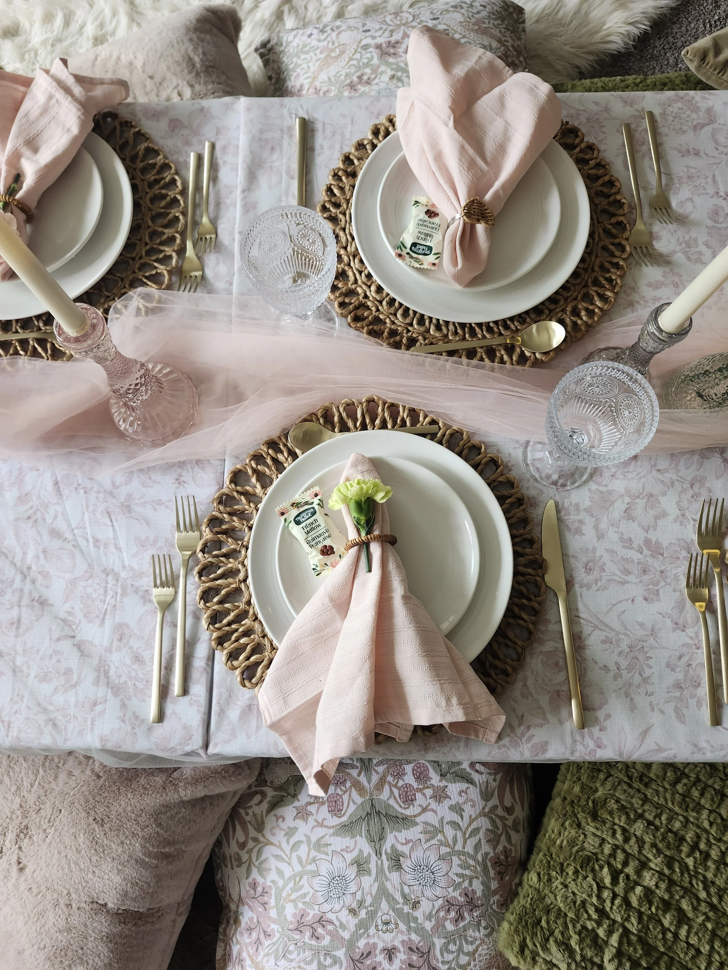 Table set for a meal with woven placemats, white plates, pink napkins, gold utensils, crystal glasses, and decorative elements, on a table with a pink and white tablecloth surrounded by various pillows and cozy blankets.