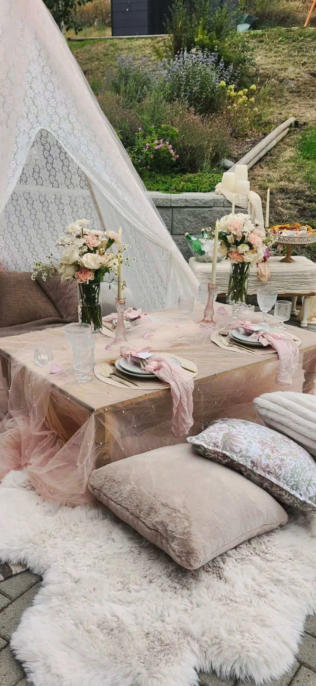 Outdoor table set for a celebration with pink and white flowers, candles, and elegant tableware, decorated with soft pink fabric and pillows, placed on a fluffy white rug.
