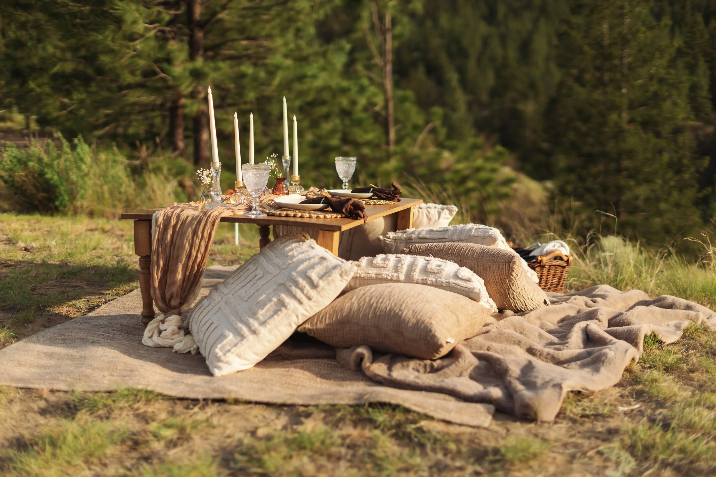 Outdoor picnic setup with a low wooden table, candles, glassware, plates, and cushions on a blanket surrounded by trees.