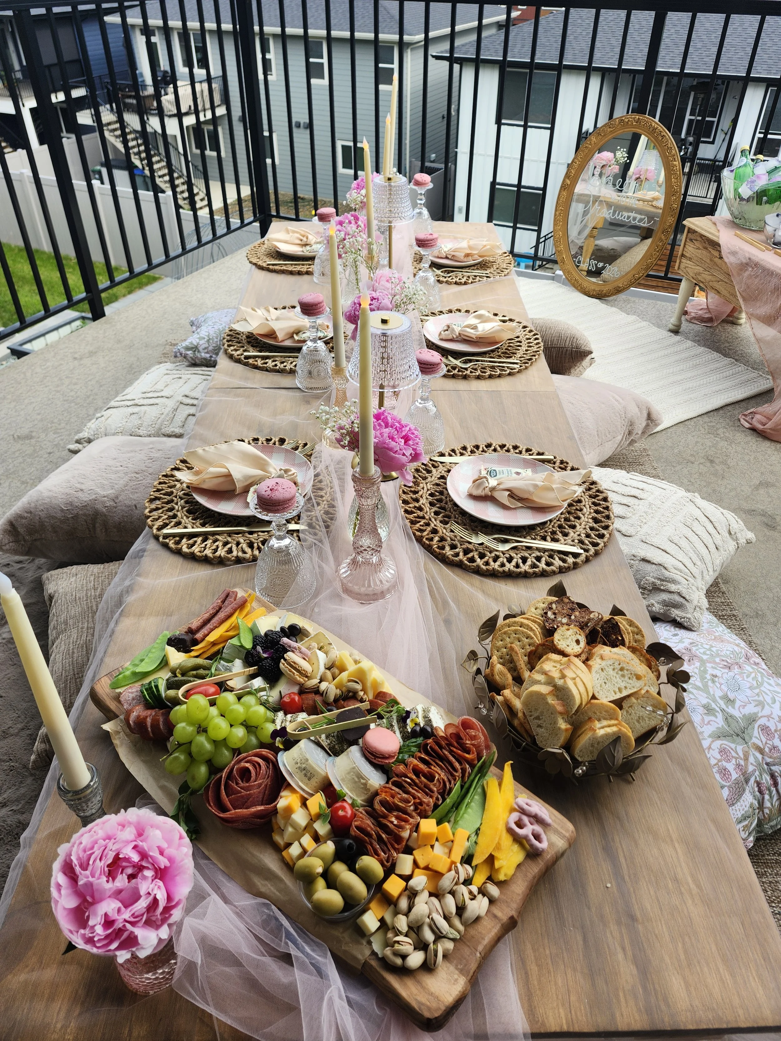 A dining table set with pink candles, pink flowers, and elegant tableware, situated on a balcony overlooking neighboring houses. The table features a large charcuterie board with grapes, cheese, meats, crackers, and sweets, and a mirror with a gold f